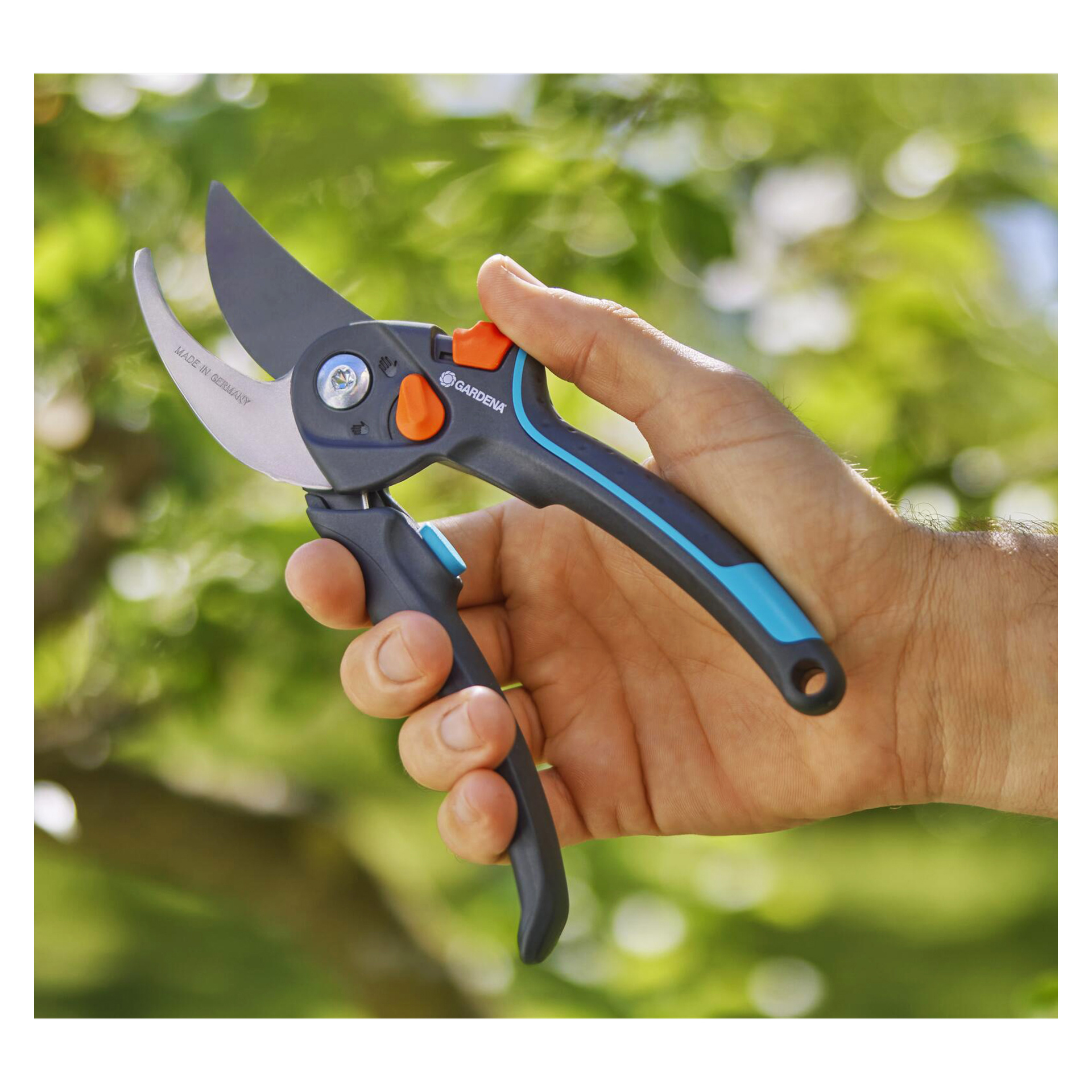 En hand håller Gardena PremiumCut pro flex beskärningssaxar, svarta och blå handtag med orange lås, utomhus mot grön suddighet.