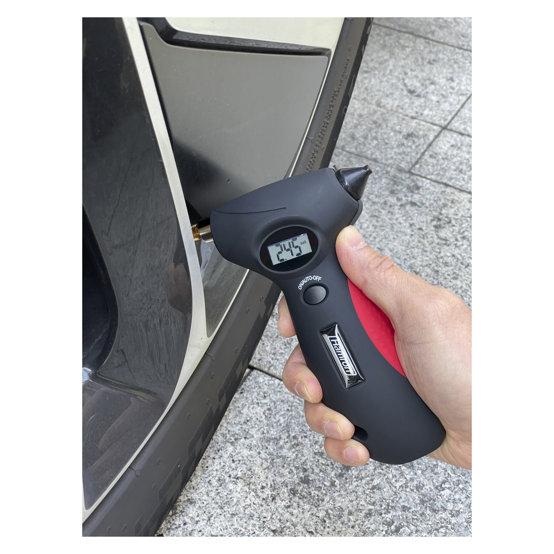 En hand kontrollerar trycket på ett bildäck med Hamron 5-i-1 nödhammare, som visar 24,5 PSI, på grå plattor.