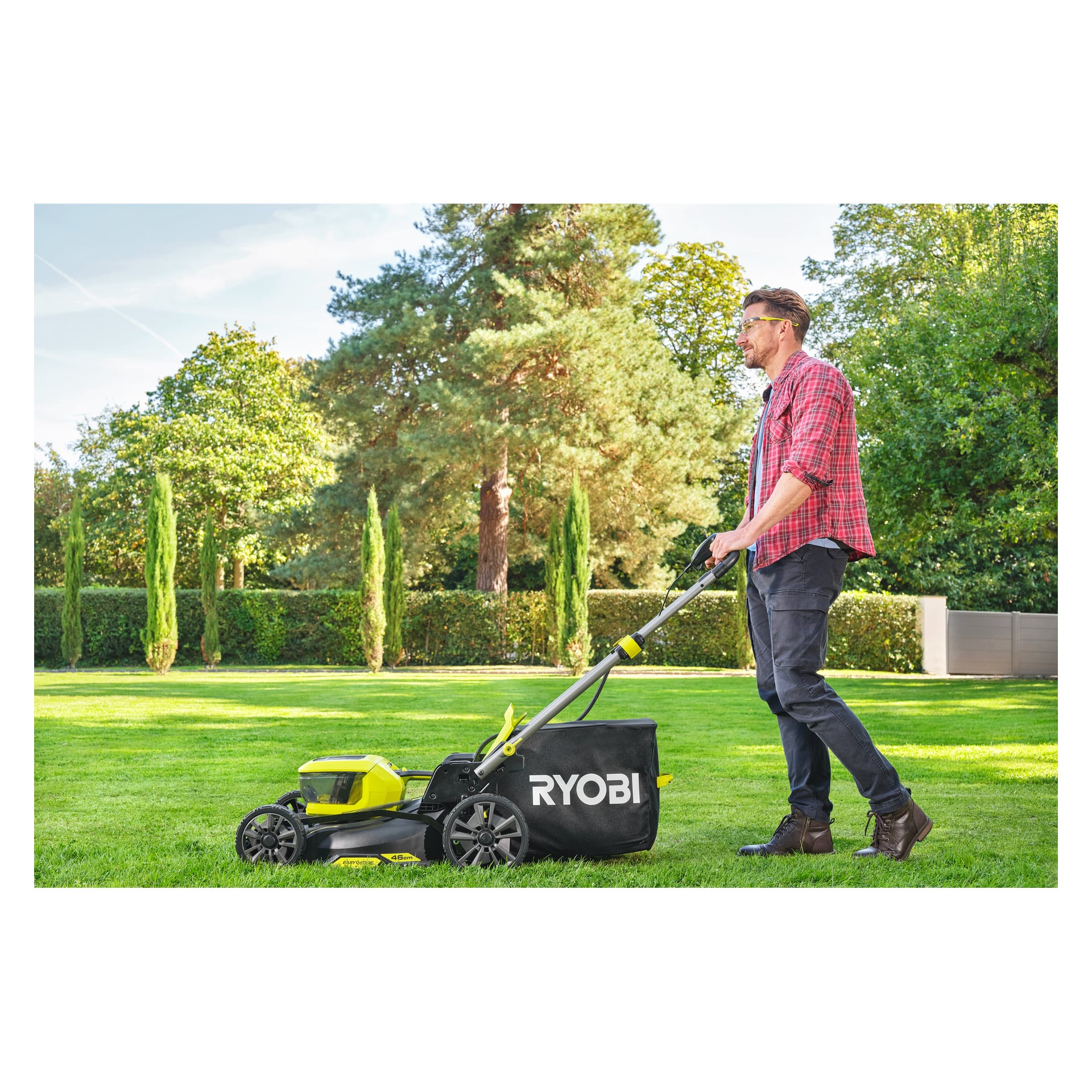 En man med skyddsglasögon och en rödrutig skjorta klipper en grön gräsmatta med en Ryobi One+ batteriklippare bland träd och häckar.
