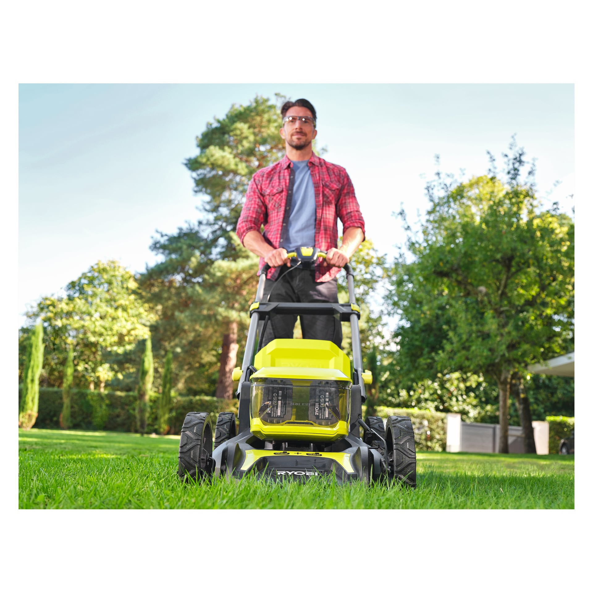 En man i en röd rutig skjorta och skyddsglasögon klipper en grön gräsmatta med en Ryobi One+ sladdlös gräsklippare, träd och en byggnad bakom.