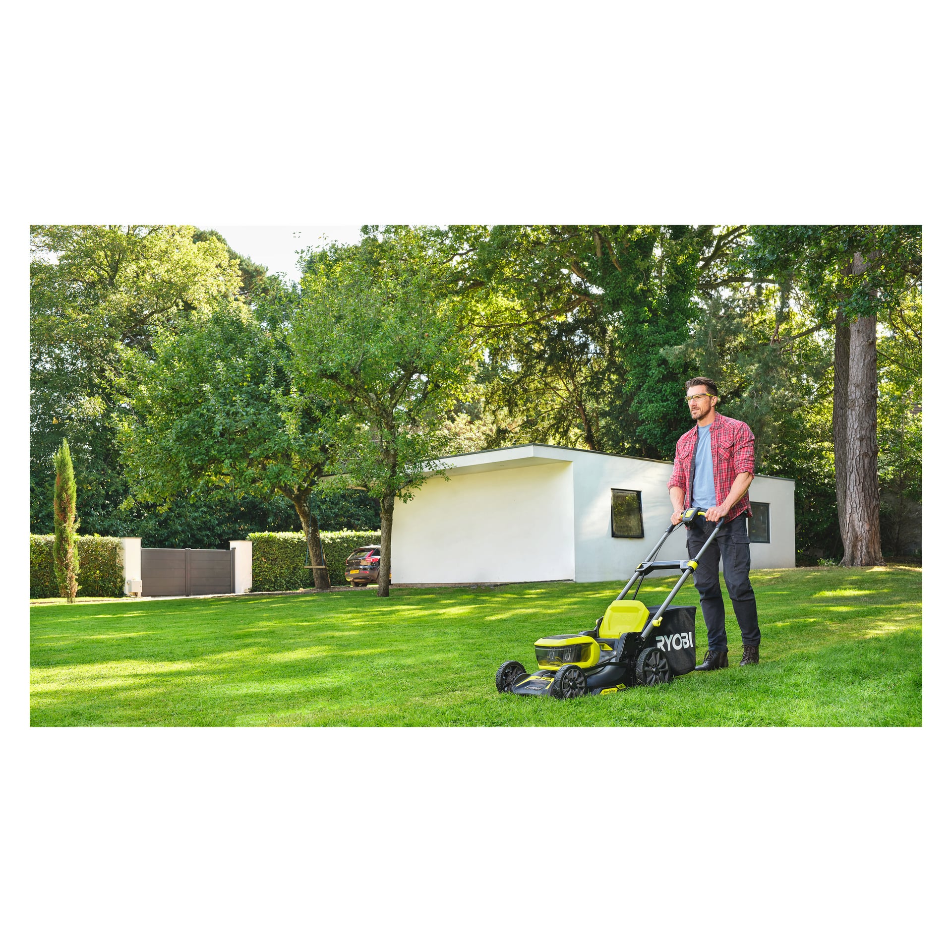 En man i rödrutig skjorta och glasögon använder en Ryobi One+ batterigräsklippare på en grön gräsmatta utanför ett modernt vitt hus med träd.