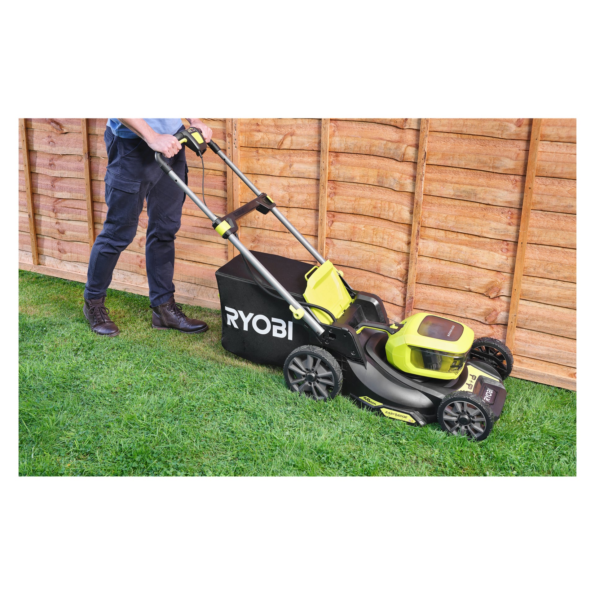 En person som klipper grönt gräs bredvid ett trästaket med en Ryobi One+ batterigräsklippare RY18LMX46A-0 med en gräslåda fäst.