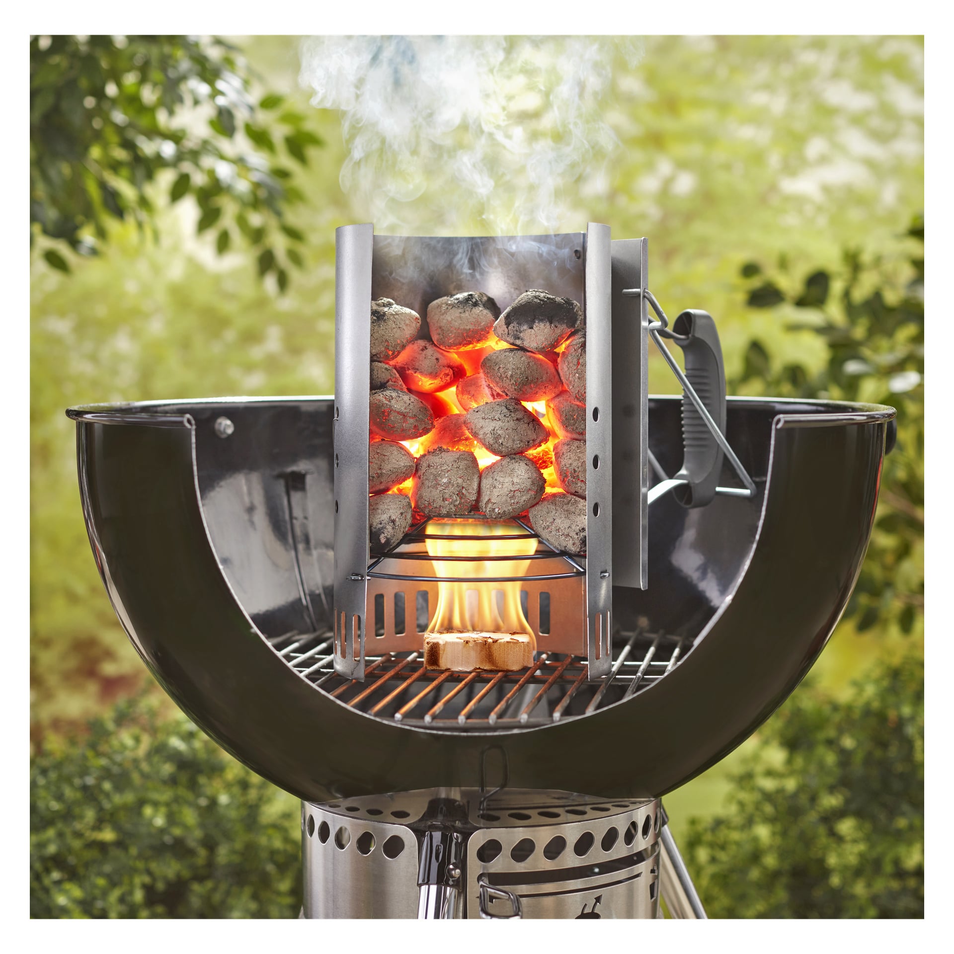 Weber Grillin briketit loistavat savupiipun starttimessa pyöreän grillin päällä, savu pyörii ja taustalla on lehtivihreä.