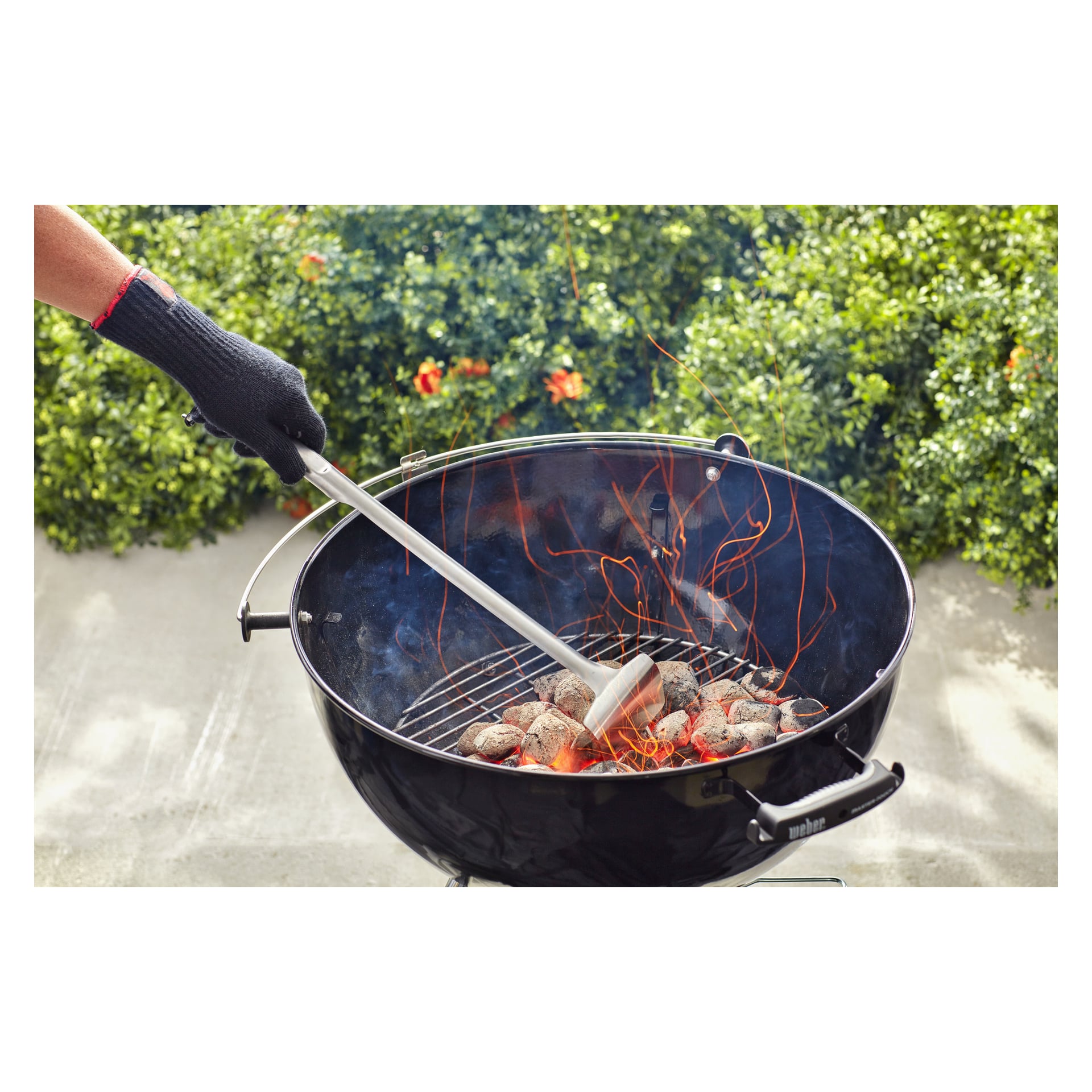 Hansikoitu käsi asettaa pihdeillä Weber Grillin briketit pyöreään grilliin, kun savu ja kipinät nousevat, ja vihreitä pensaita on jäljellä.