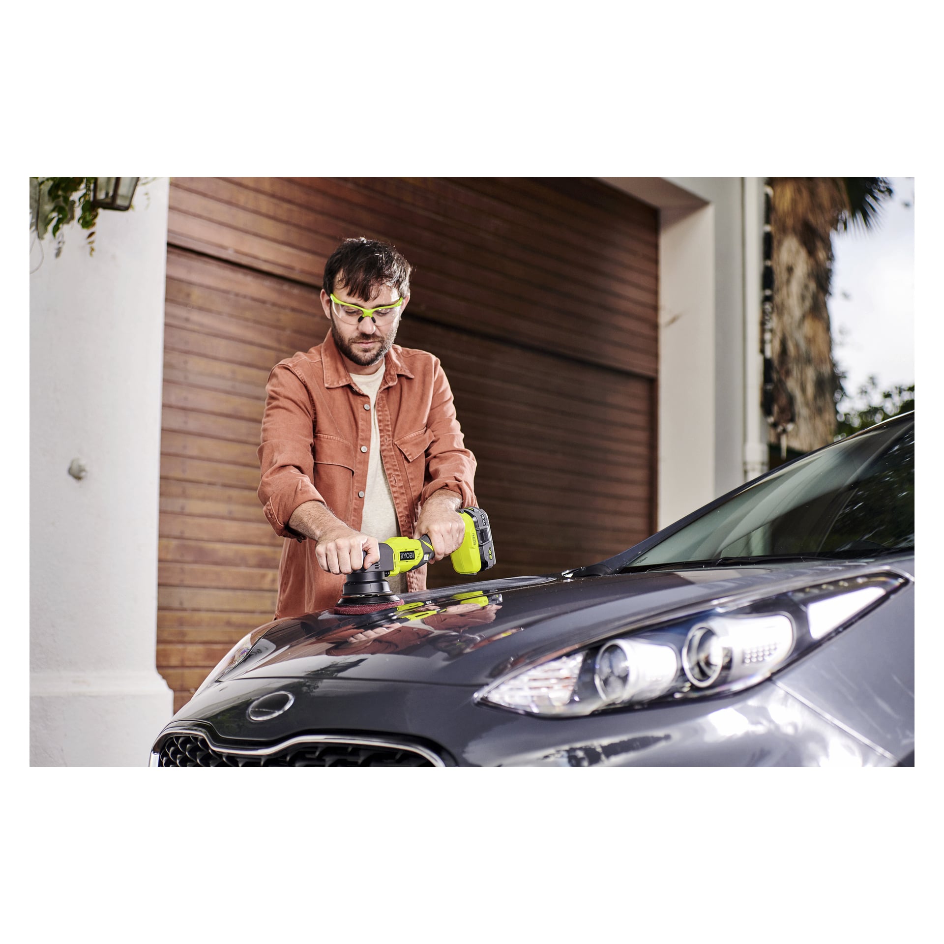 En man med skyddsglasögon och brun skjorta putsar en mörkgrå bil med en Ryobi R18P-140 nära en garageport i trä.