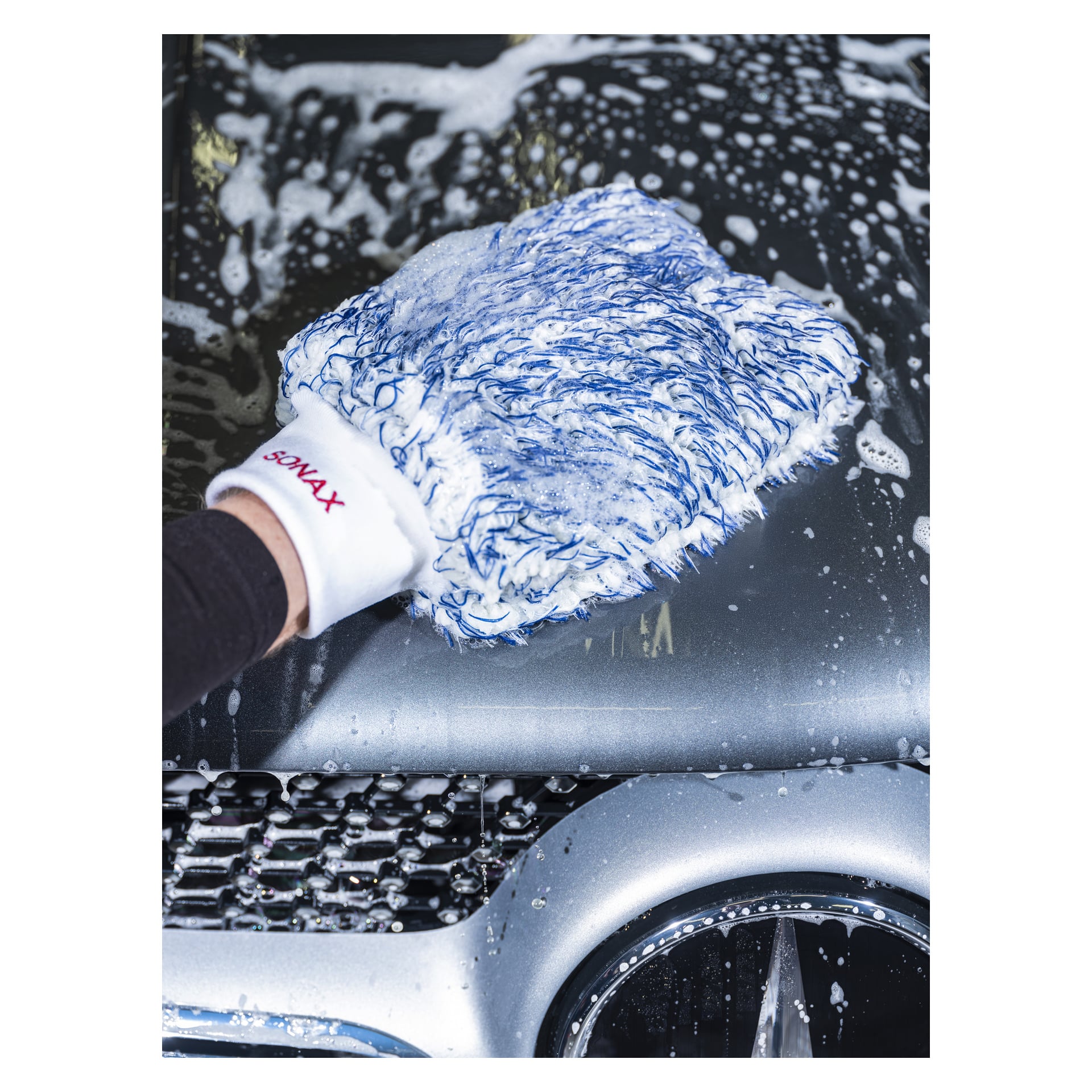 En hand i Sonax Wash-handsken Xtreme Wonder Wash Glove, inbäddad med tvål, torkar av huven på en silverfärgad bil. Skum och vatten är utspridda över ytan, och en del av grillen är synlig.