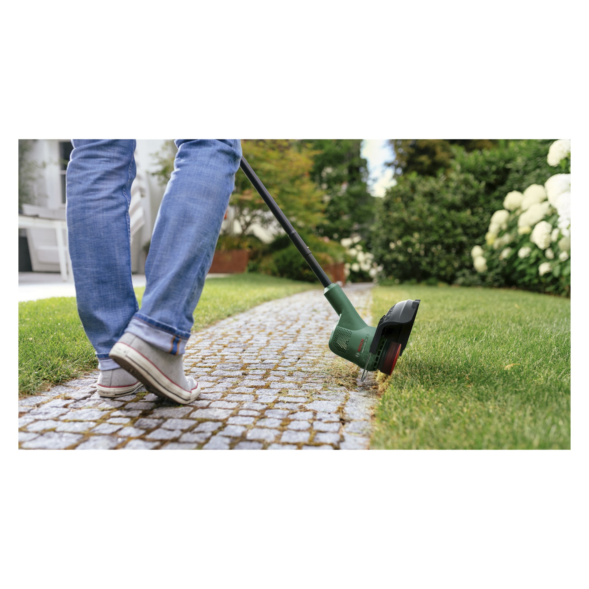 En person i jeans och sneakers kantar en trädgårdsgräsmatta vid en stenstig med hjälp av en Bosch Power 4 All Moder och trimmer.