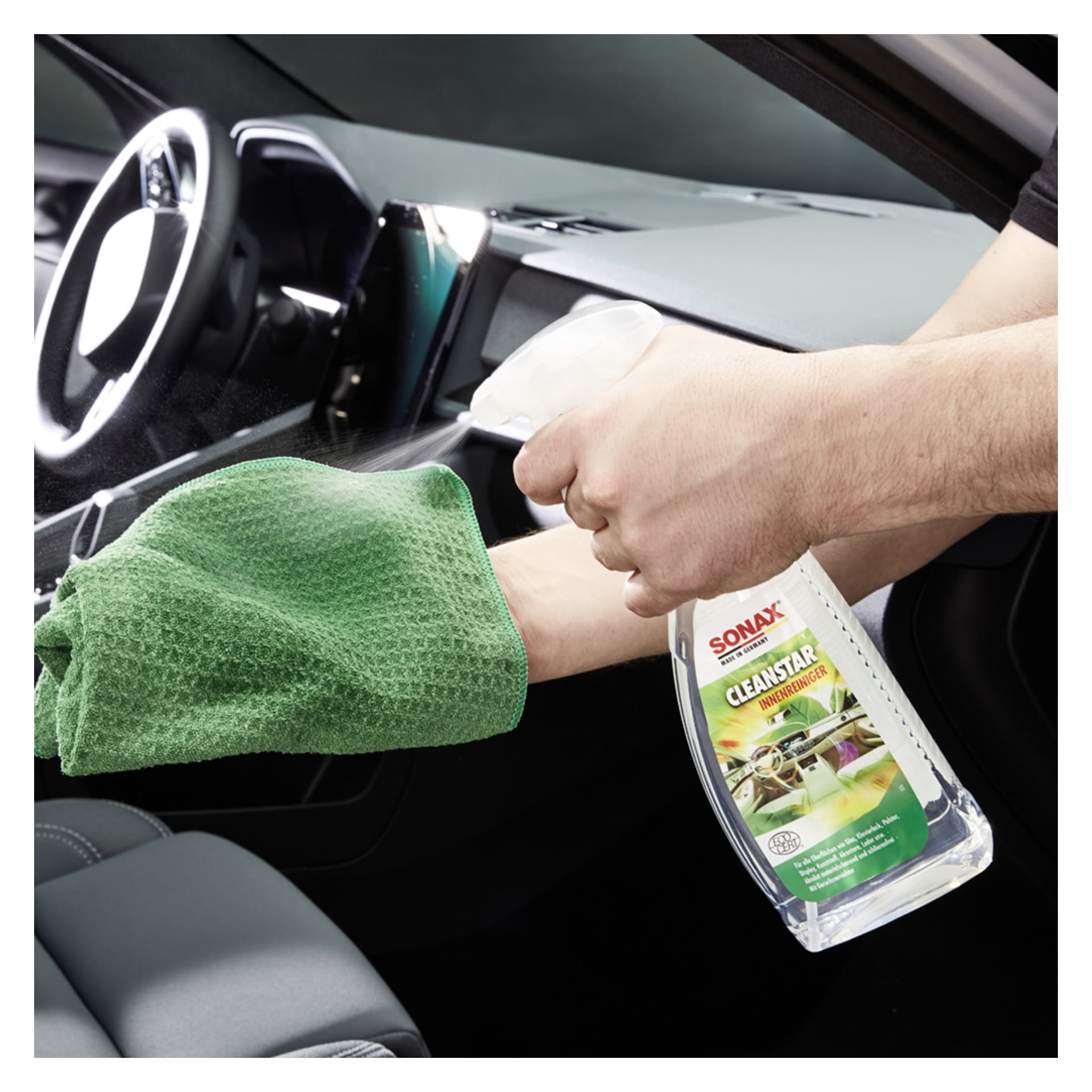 Man applicerar Sonax Interior Cleaner CleanStar APC på en grön mikrofiberduk och förbereder sig för att torka av bilens instrumentbräda.