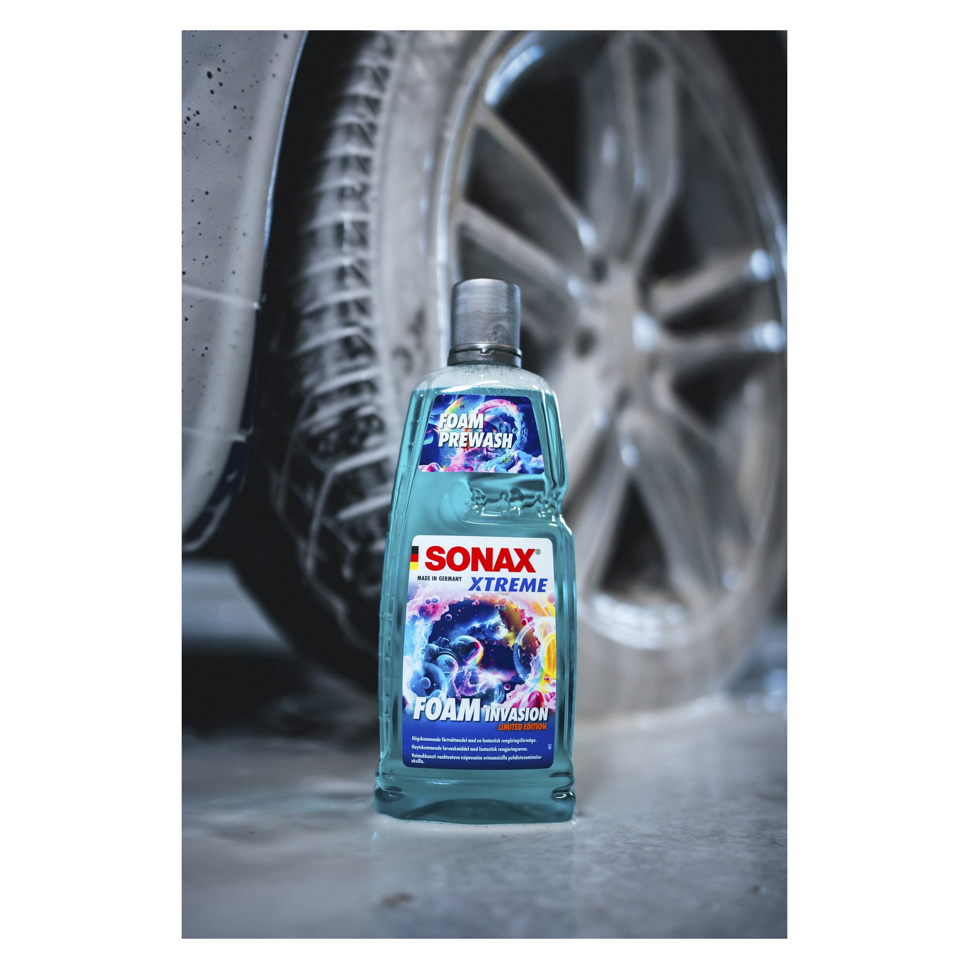 En flaska med texten "Car washing shampoo Xtreme Foam Invasion Limited Edition by Sonax" står på marken framför bilens hjul och däck, med synlig smuts på karossen.