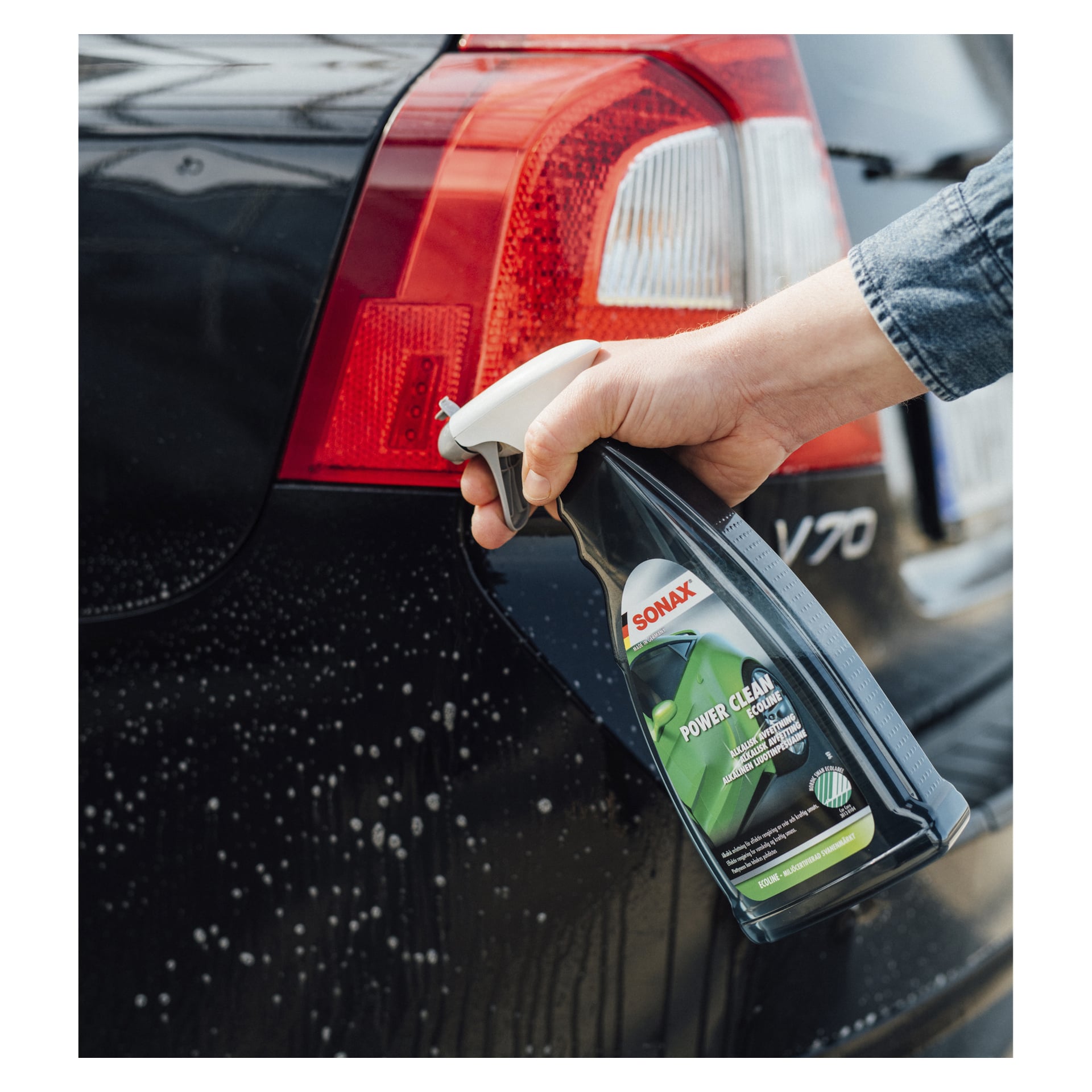 En handsprayad Sonax Alkaline Degreaser Power Clean Ecoline på den våta, tvåliga bakdelen av en svart bil, vilket visar dess baklykta och V70-emblem.