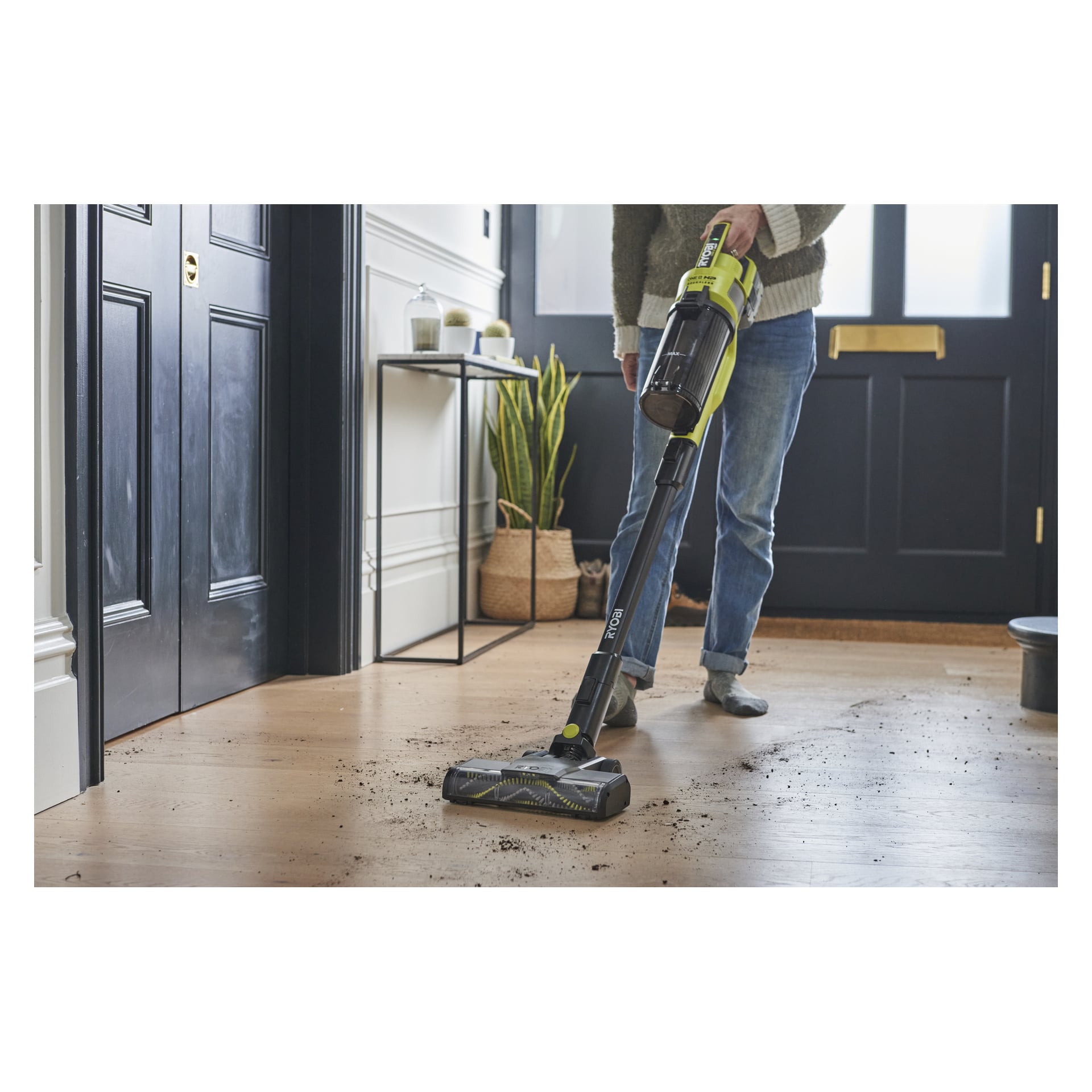 Någon rengör smuts från ett trägolv nära ingången med en Ryobi One+ RSV18X-1X40 dammsugare, med växter och dekorationer längs väggen.