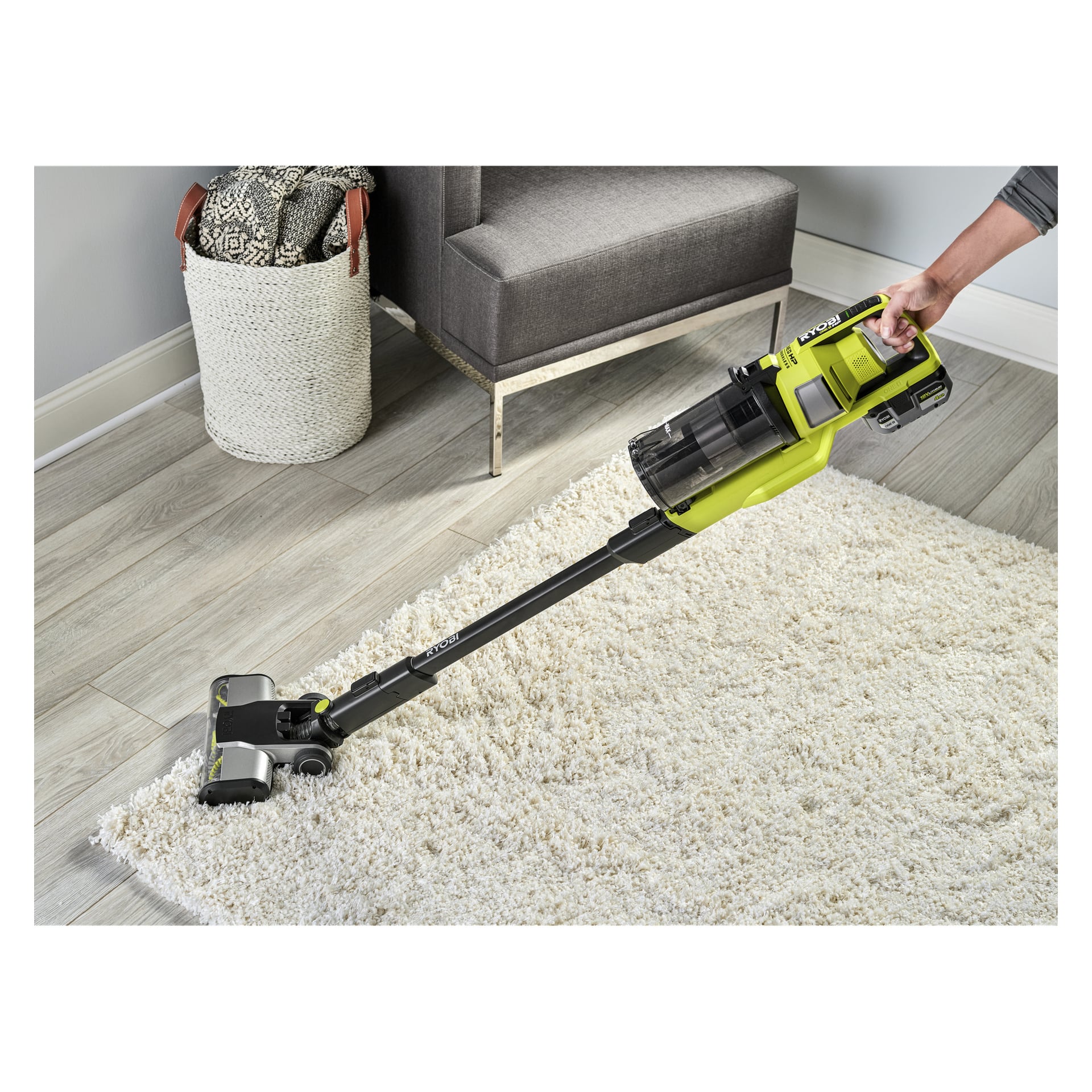 Någon dammsuger en vit matta i ett modernt vardagsrum med Ryobi One+ Upright RSV18X-1X40, nära en grå stol och korg.
