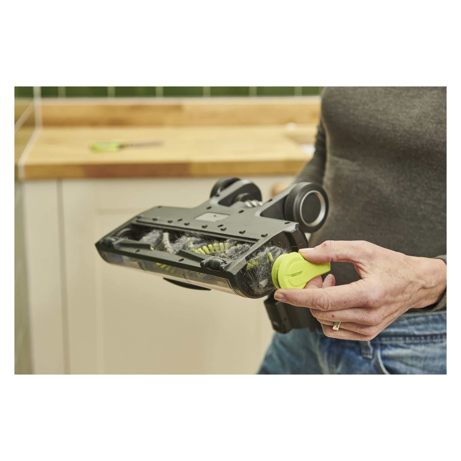 Någon håller i en Ryobi One+ Upright dammsugare RSV18X-1X40 och tar bort en gul del från borsthuvudet nära en köksbänk med gröna plattor.