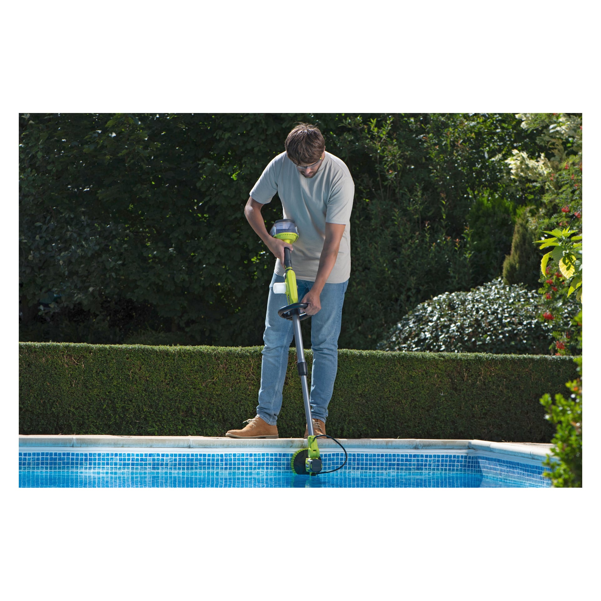 Någon som bär jeans, en lätt skjorta och stövlar använder Ryobi One+ Cleaning-borsten vid en pool, med häckar och växter bakom.