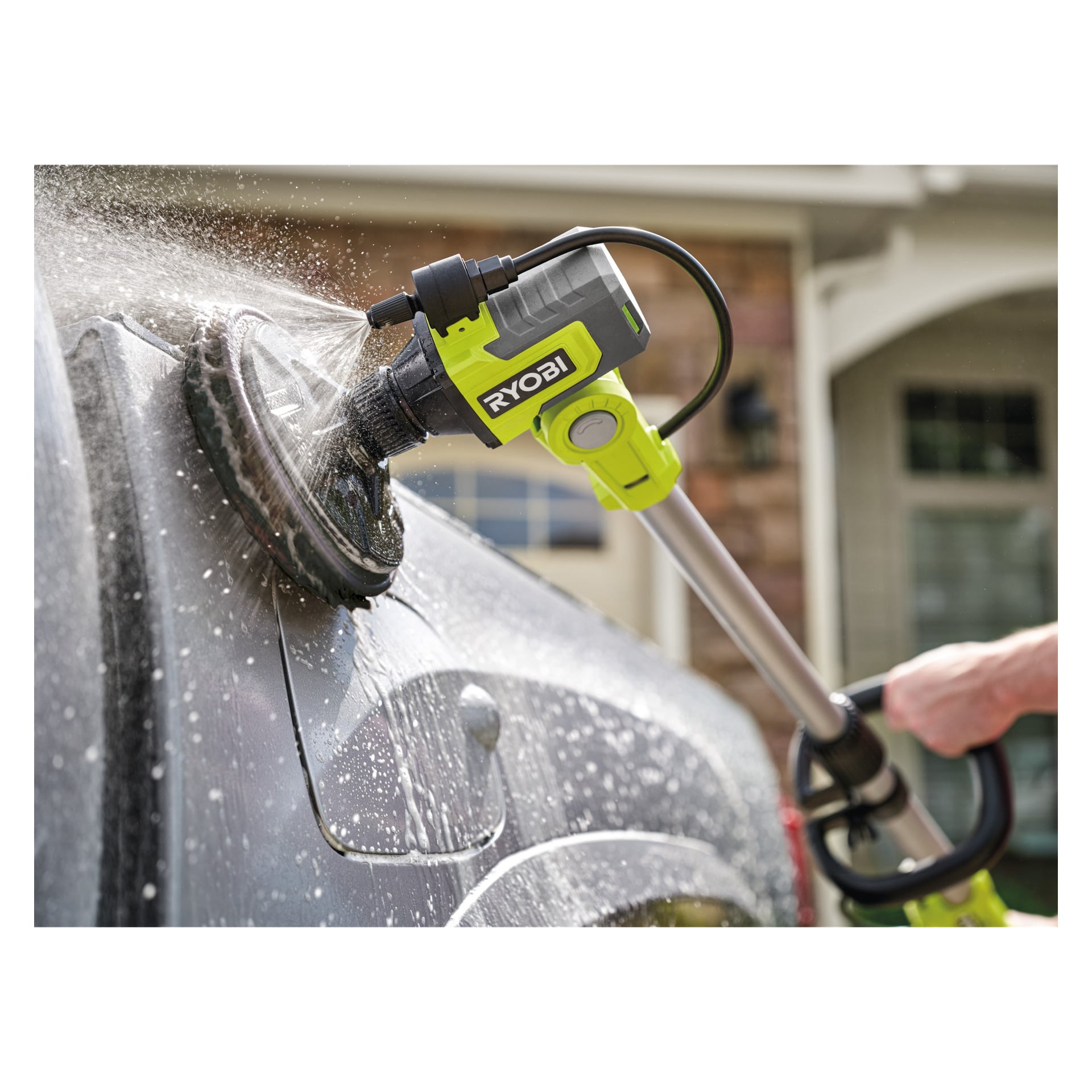 Någon rengör en grå bils sidospegel med en Ryobi One+ Cleaning-borste, vatten sprutar medan den snurrar. Ett hus syns bakom.