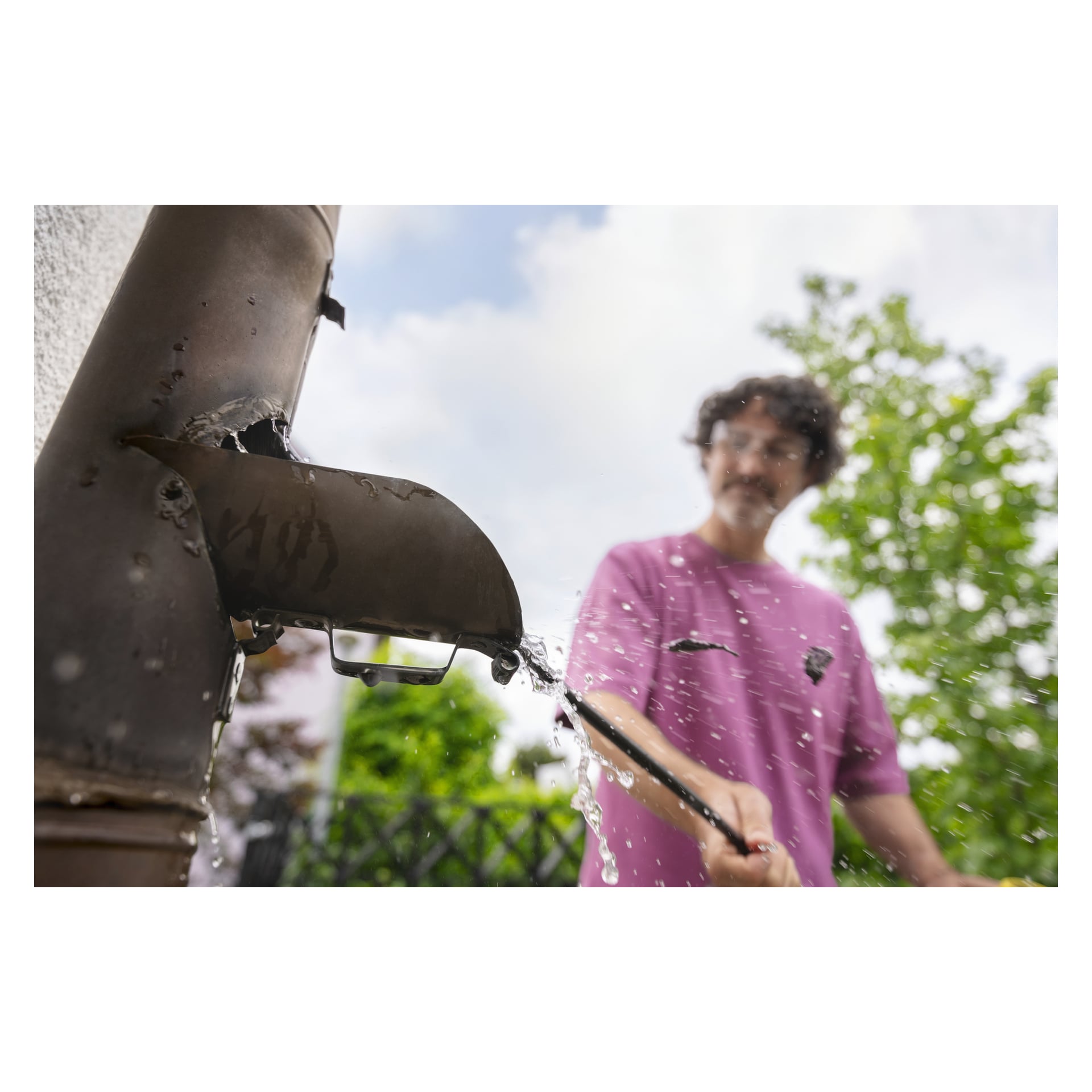 Iklädd en rosa skjorta rengör någon en utomhus metallränna med Kärcher Pipe Cleaning Kit PC 15; Vatten skvätter i närheten.