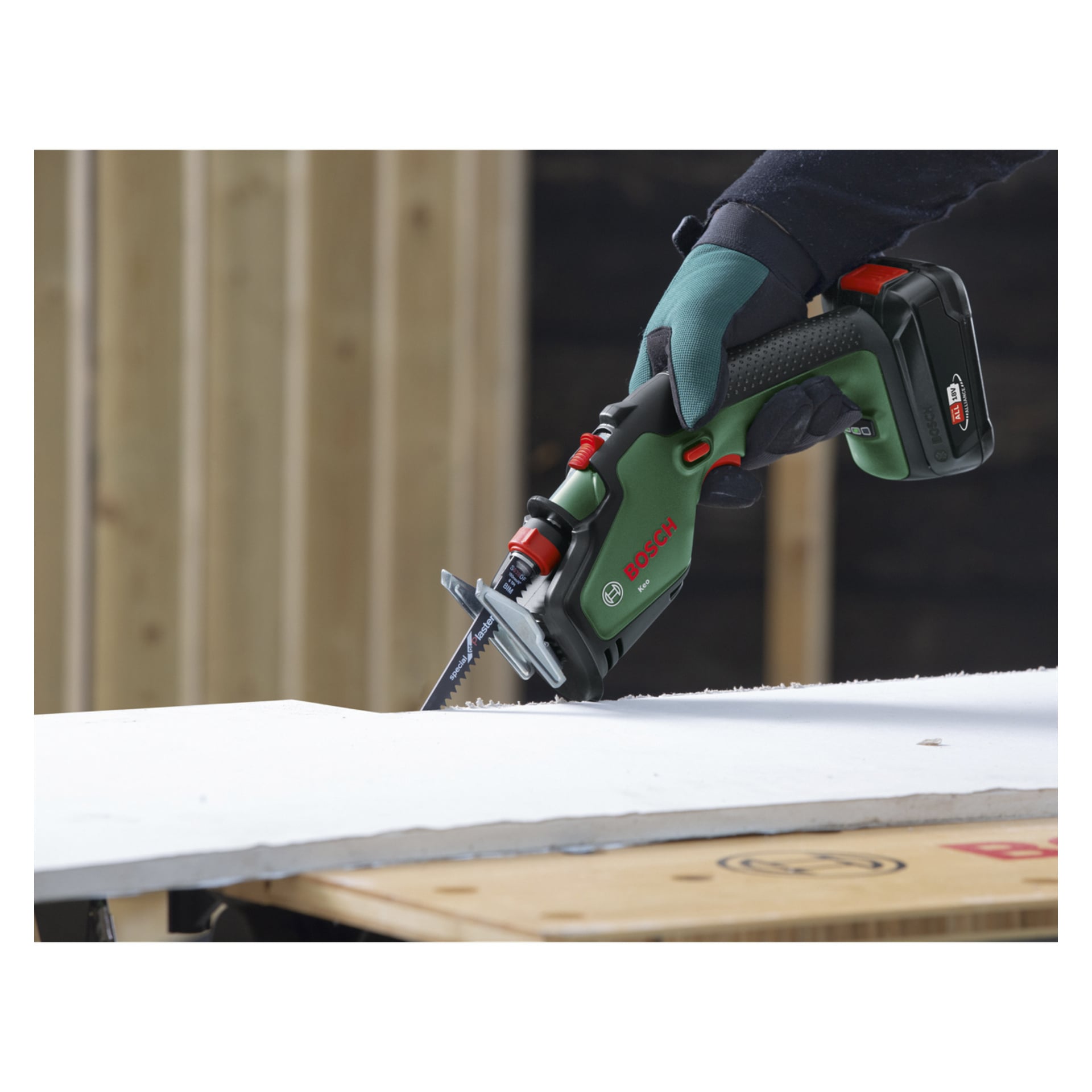 Handskbeklädda händer använder en Bosch Power 4 All Keo beskärningssåg för att skära en vit skiva, troligen gipsskiva, på en arbetsbänk med träbalkar bakom.