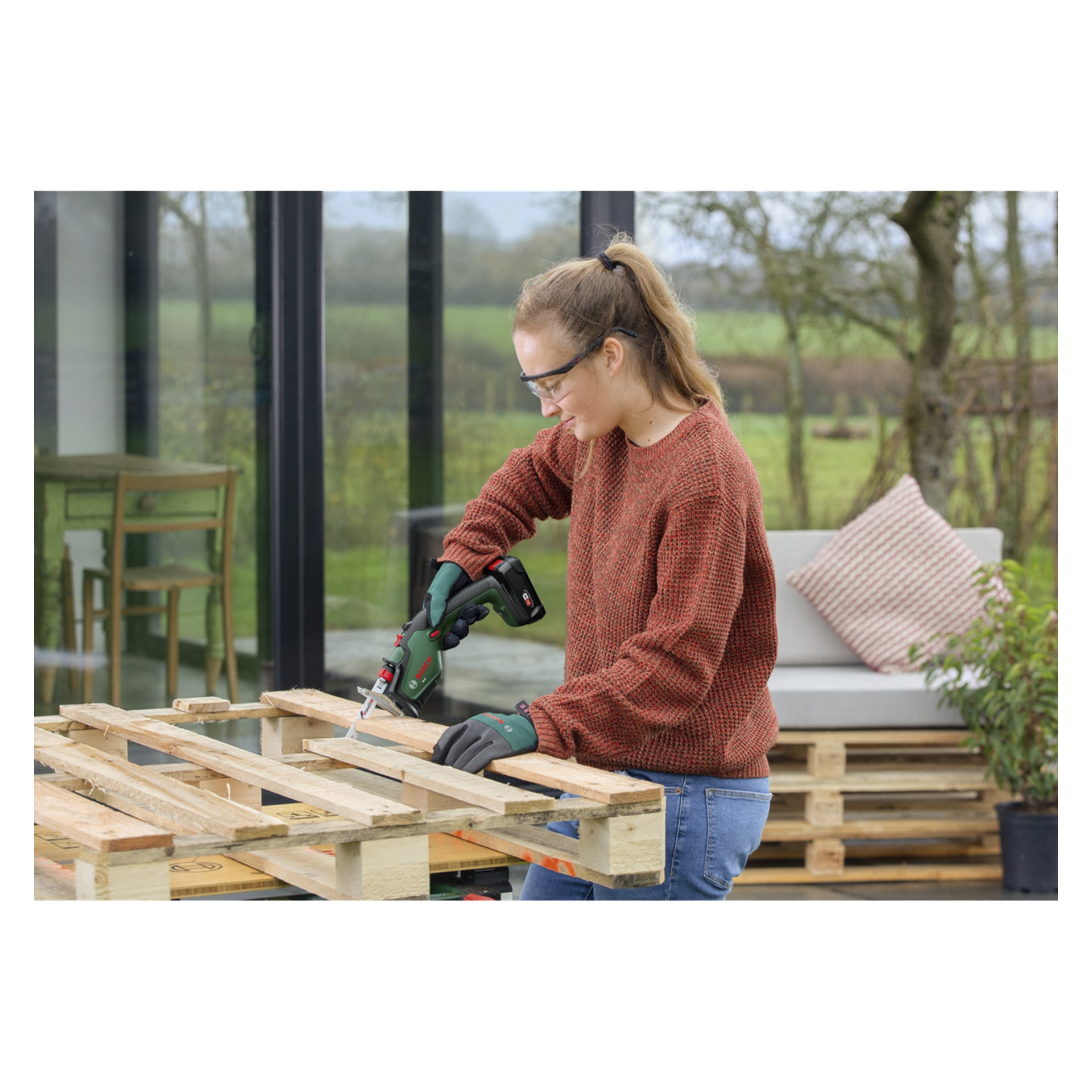 En person med skyddsglasögon och handskar använder en Bosch Keo-såg för att plocka isär en träpall utanför en modern byggnad med stora fönster.