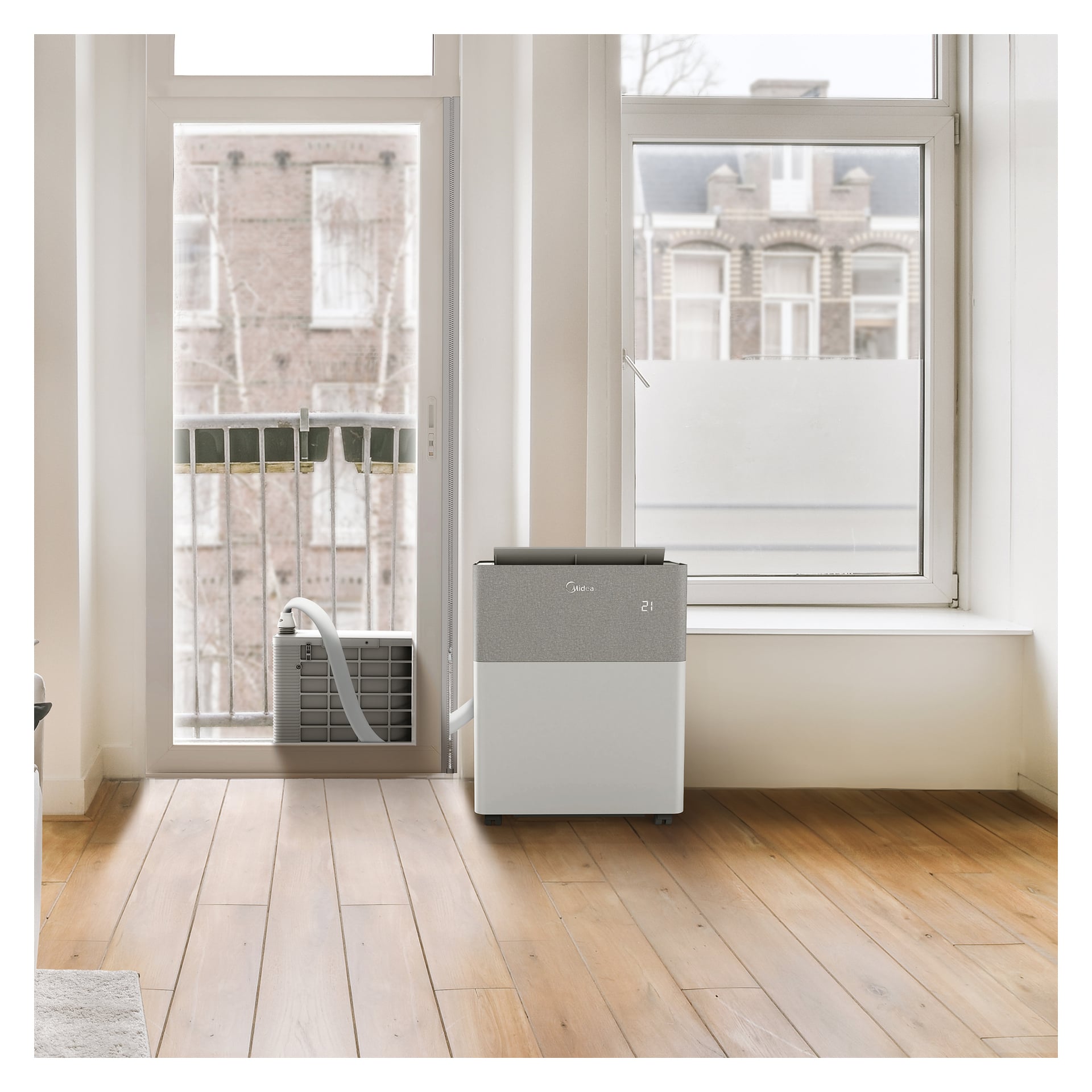 Midea PortaSplit portabel AC med värmepump står vid ett fönster på trägolv, slang ansluten till en avgaspanel; byggnader utanför.