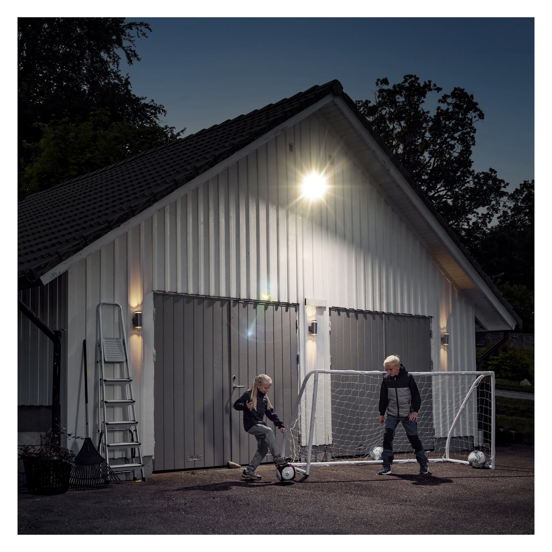 To barn spiller fotball i skumringen ved en hvit garasje under et Anslut LED-flomlys, med et lite mål, baller, stige og trær i nærheten.
