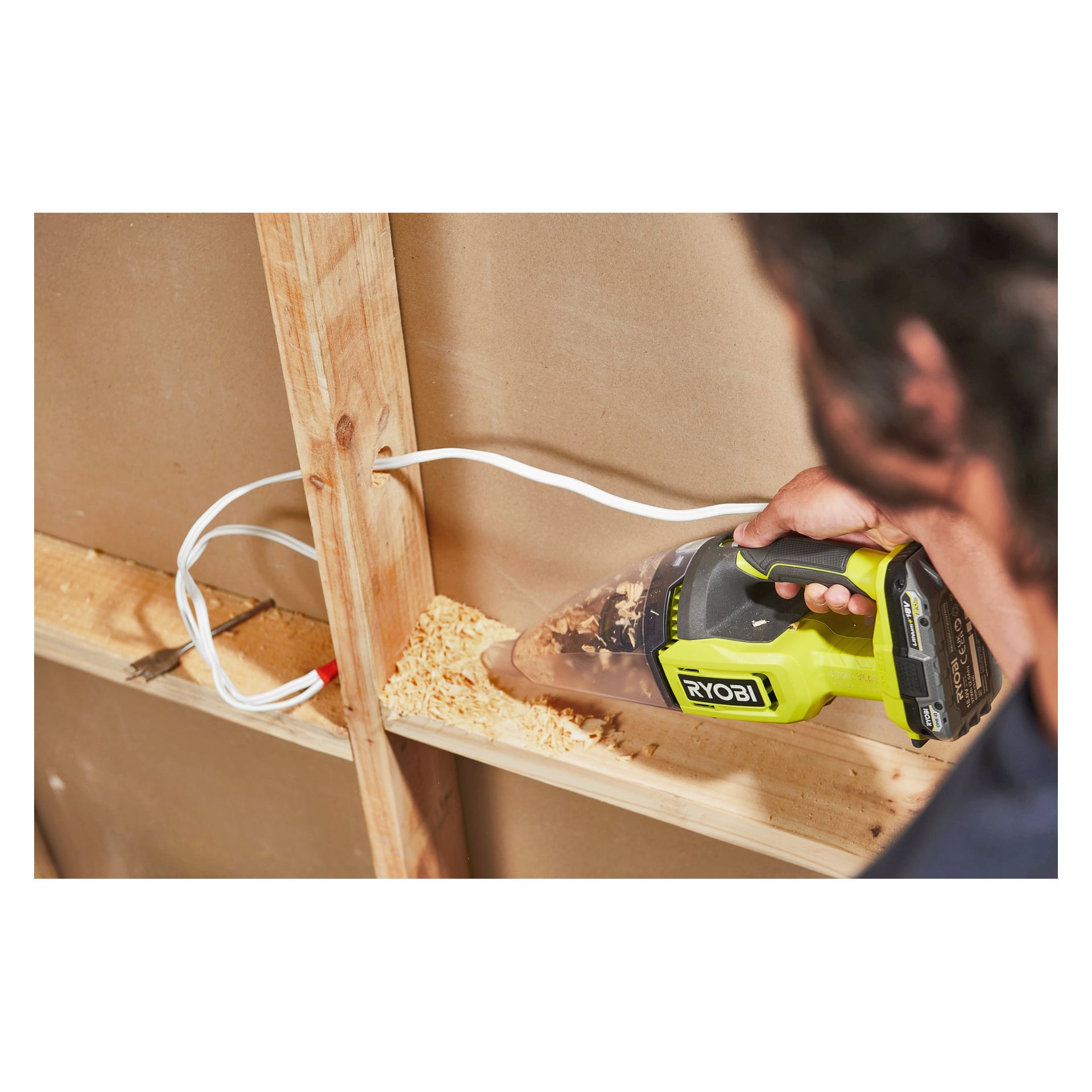 En person dammsuger träspån från en trähylla med en grön Ryobi-handdammsugare nära en vit väggkabel.