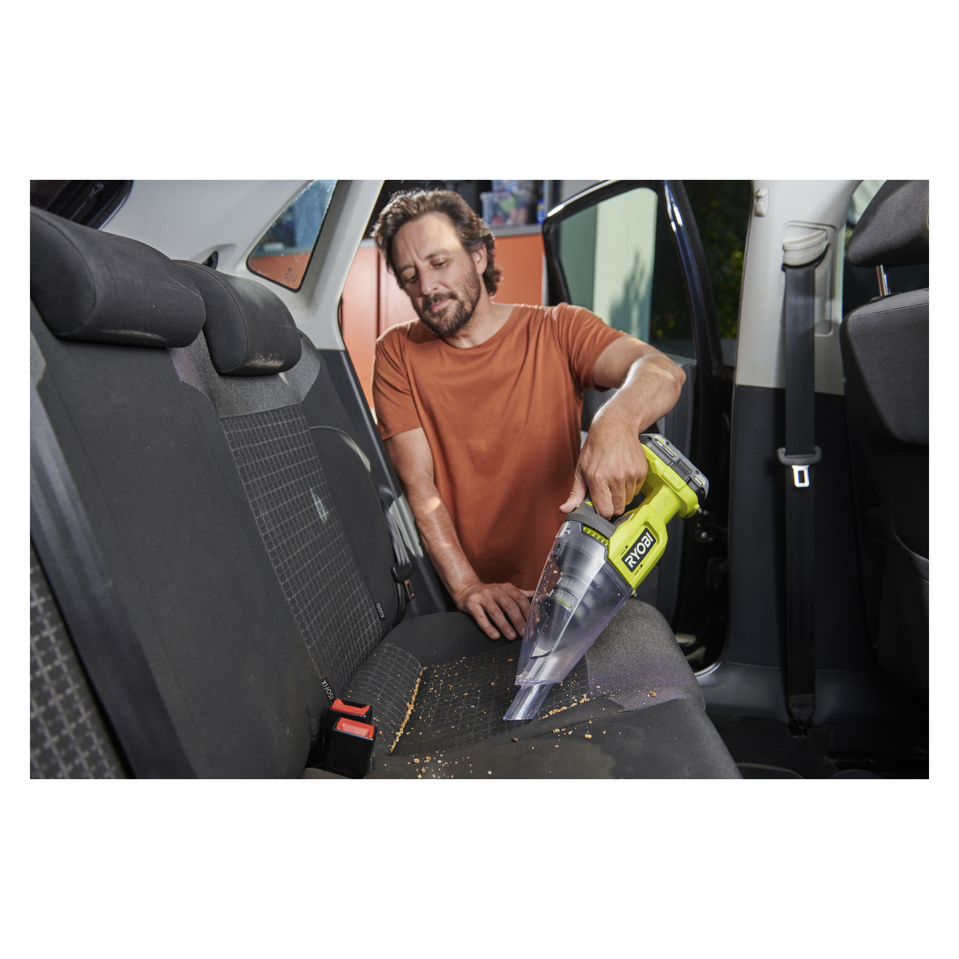 Iført en oransje skjorte støvsuger en mann smuler fra baksetet på en bil med en Ryobi One+ støvsuger; Batteriet sitter på setet.