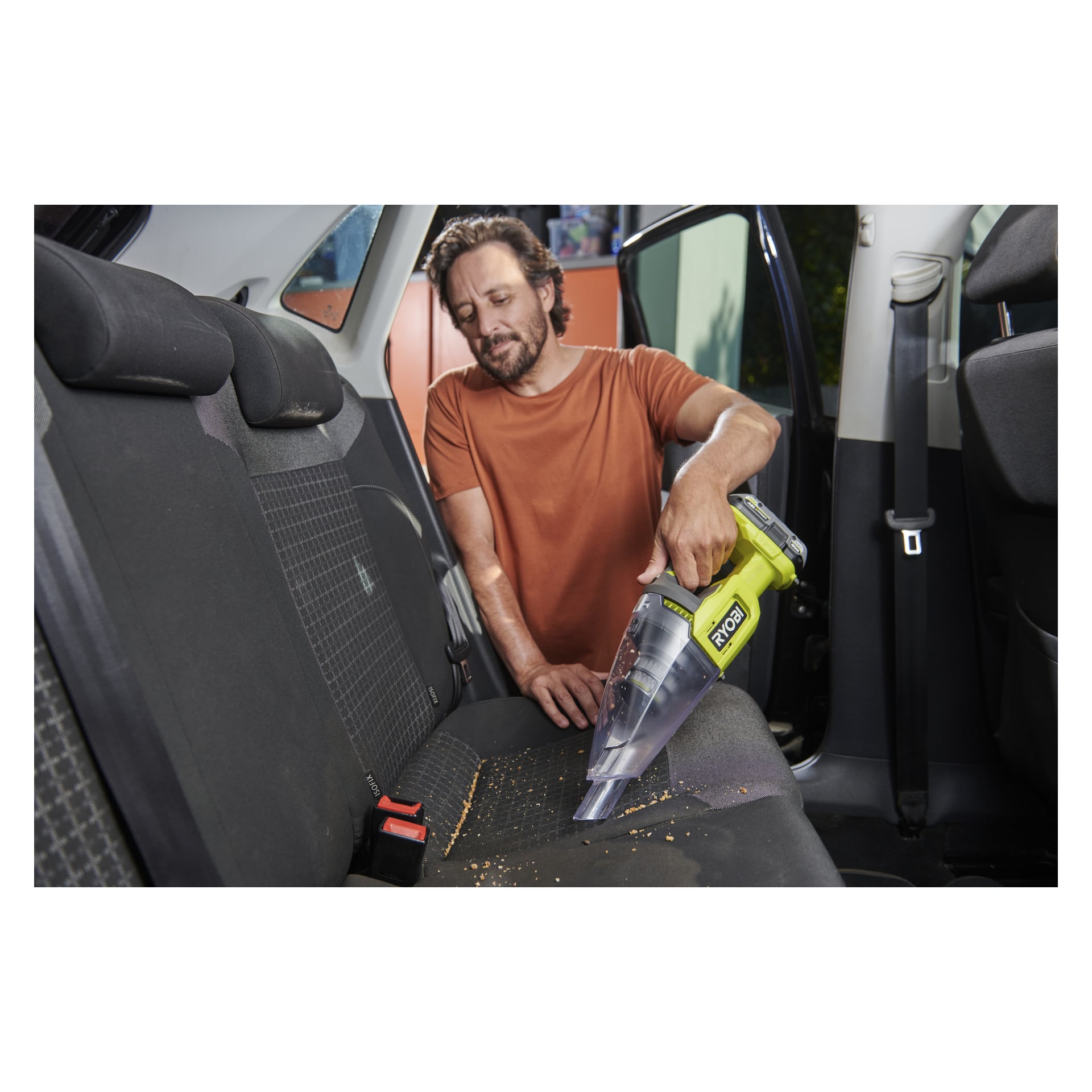 Iklädd en orange skjorta dammsuger en man smulor från baksätet i en bil med en Ryobi One+ dammsugare; Batteriet ligger på sätet.