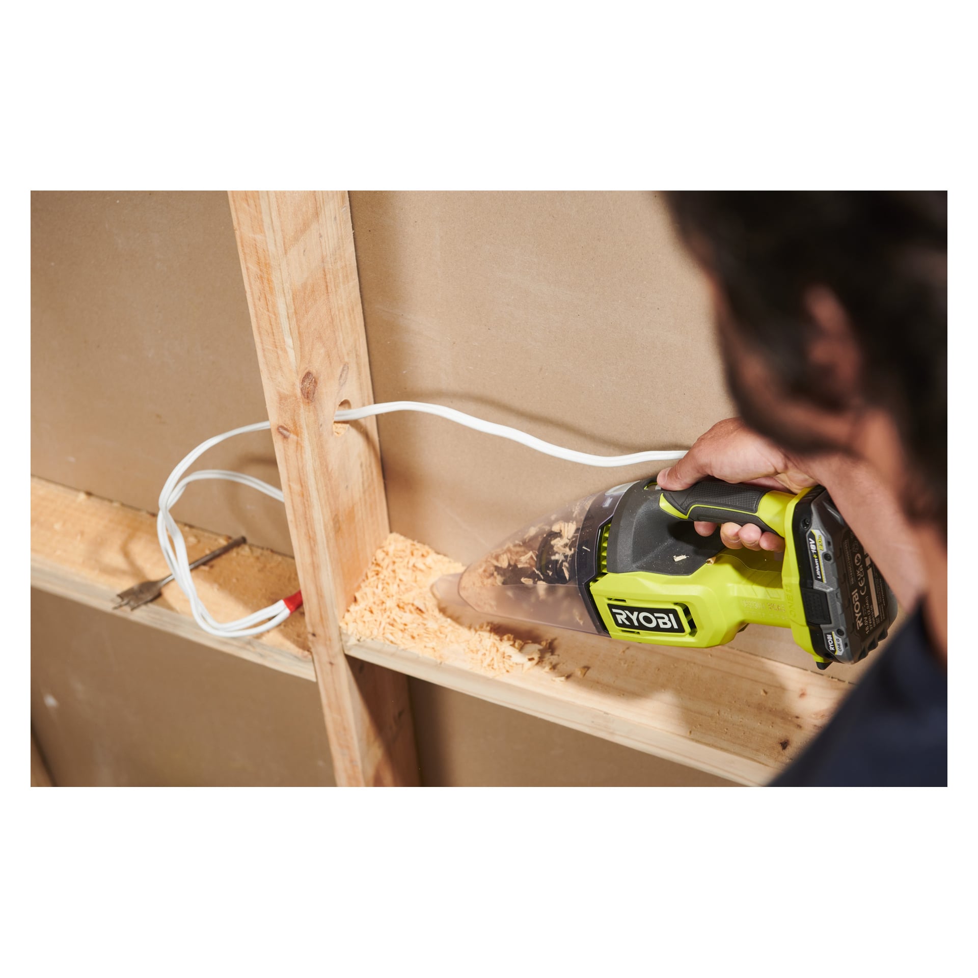 En person dammsuger träspån från en trähylla med en Ryobi One+ handhållen dammsugare, med en vit kabel längs väggen.