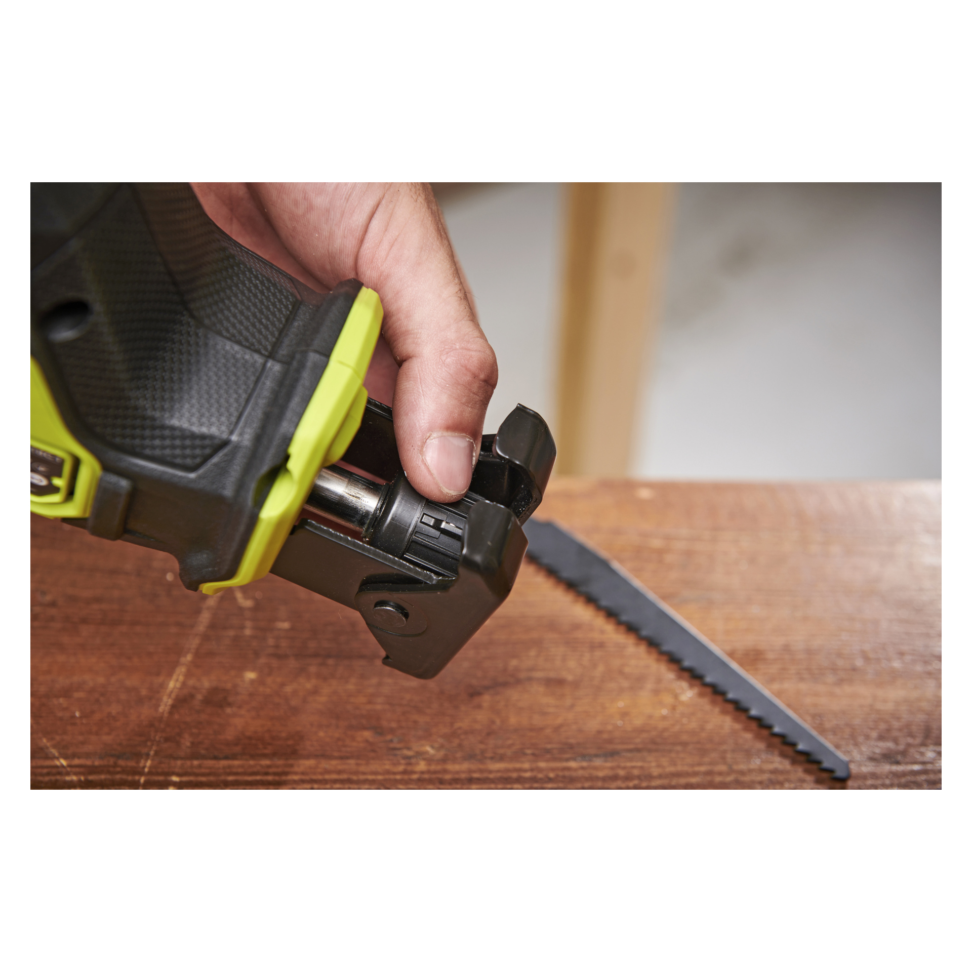 En hand för in ett blad i en Ryobi One+ HP reciprocatingsåg RRS18C1, med en träyta och ett reservblad bredvid.