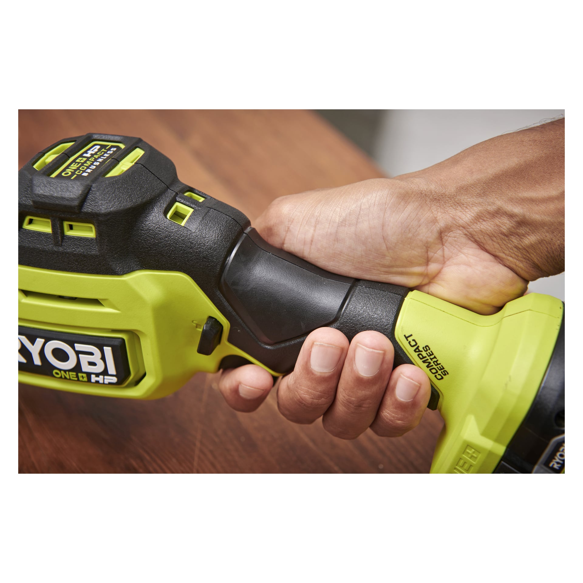 En hand greppar stadigt handtaget på en grön och svart Ryobi One+ HP Compact Series bajsåg på en träyta.