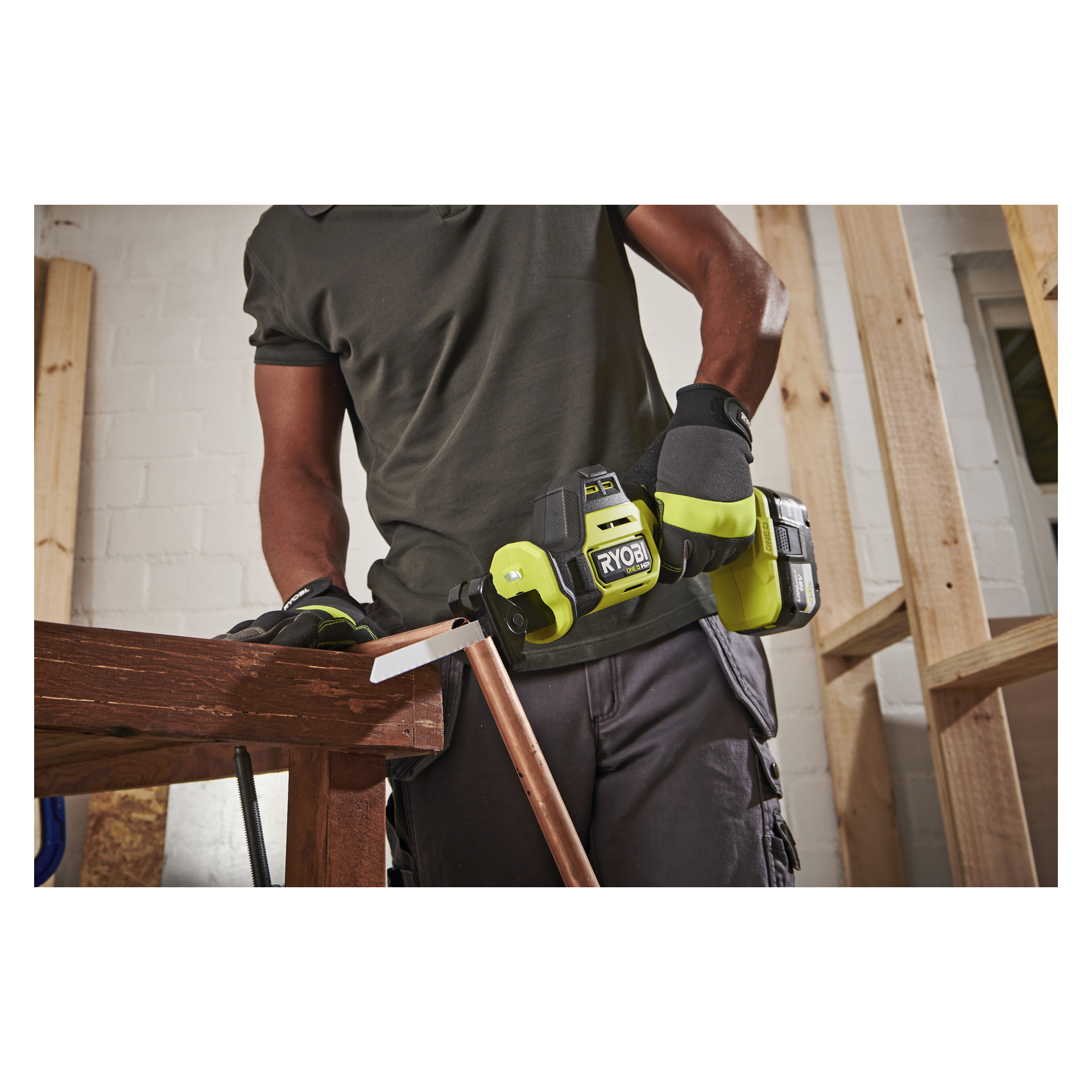 En person med handske använder en Ryobi One+ HP-såg för att kapa ett kopparrör som är fäst vid en träarbetsbänk i en verkstad.