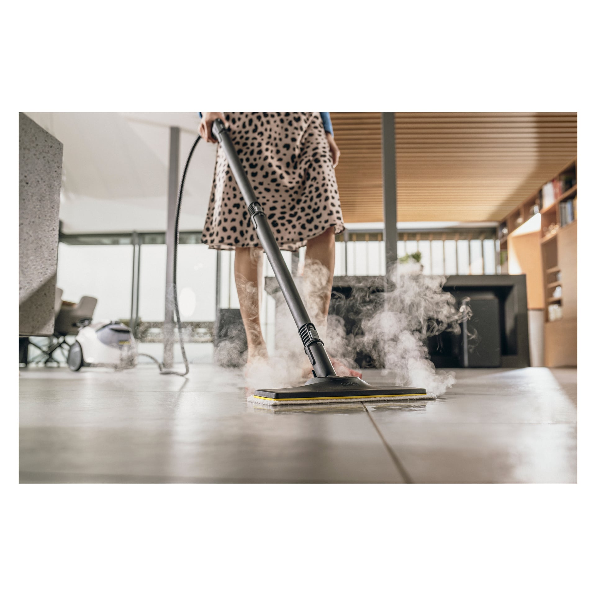 En person med mønsterskjørt bruker en Kärcher SC 5 EasyFix Iron damprenser på et flislagt gulv, med synlig damp som stiger opp mens de rengjør.