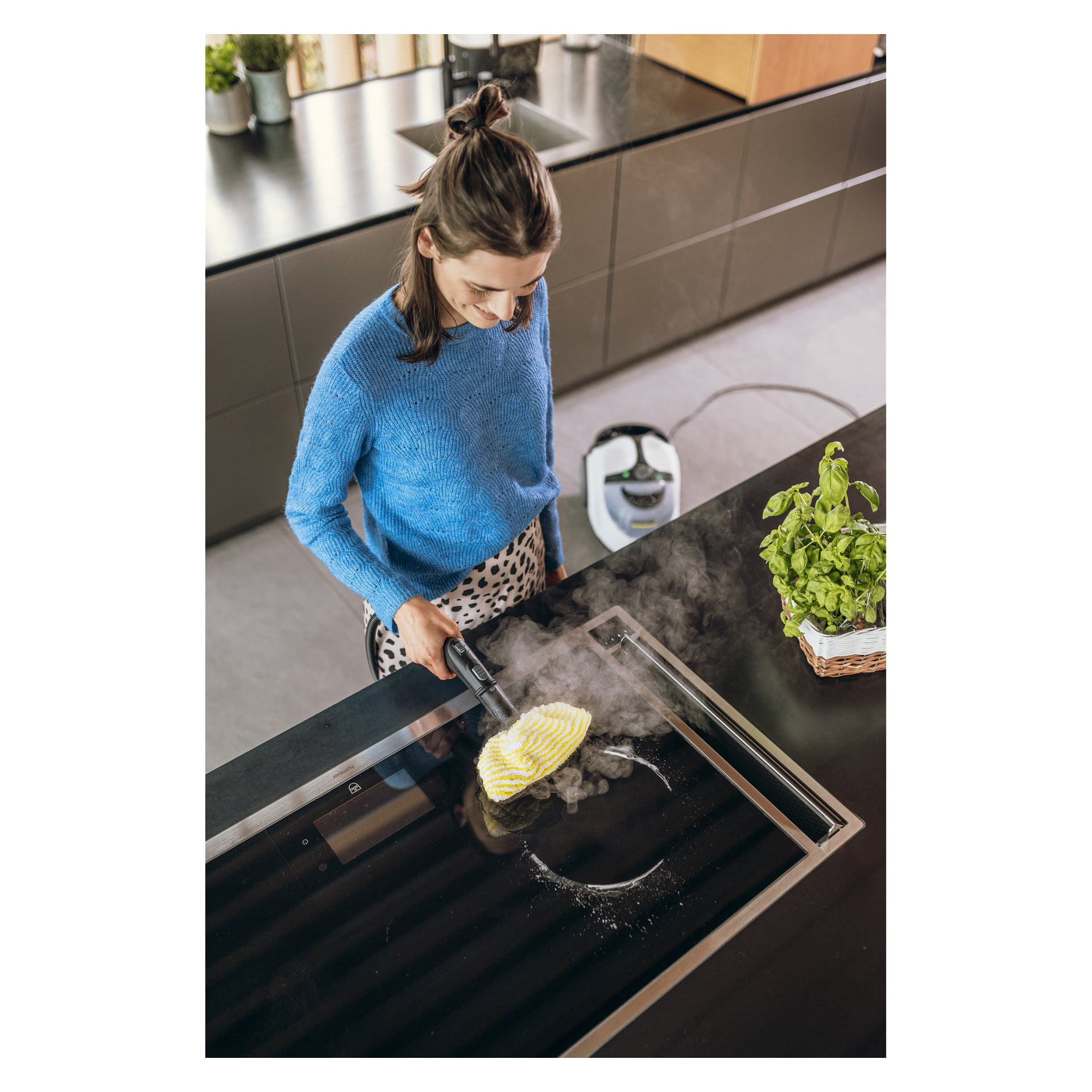 En kvinne i blå genser rengjør en svart glasskokeplate med en Kärcher damprenser på et moderne kjøkken; Basil sitter i nærheten.