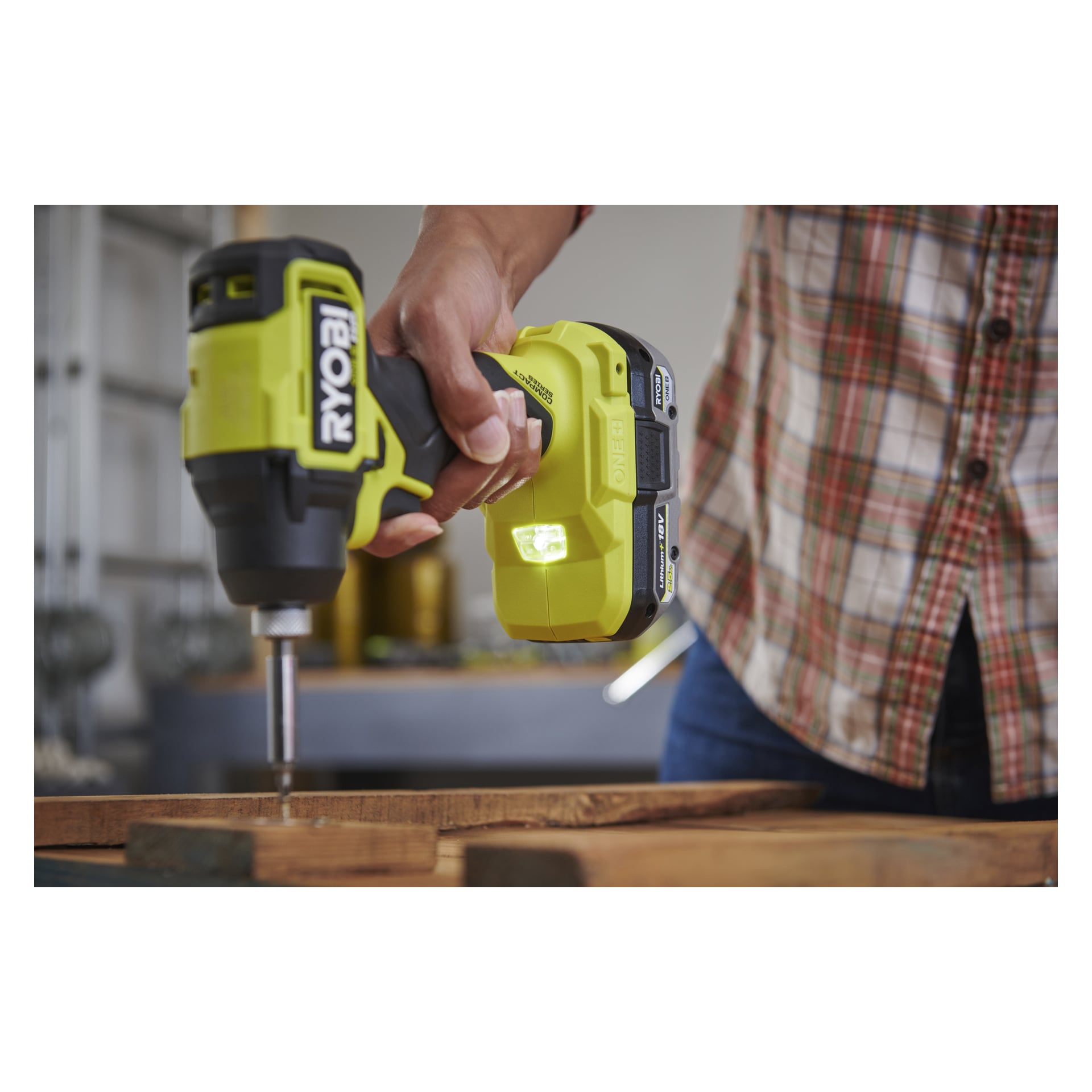 En person i rutig skjorta borrar i trä med en gul och svart Ryobi One+ HP borstlös slagdrivare, vars ljus lyser upp området.