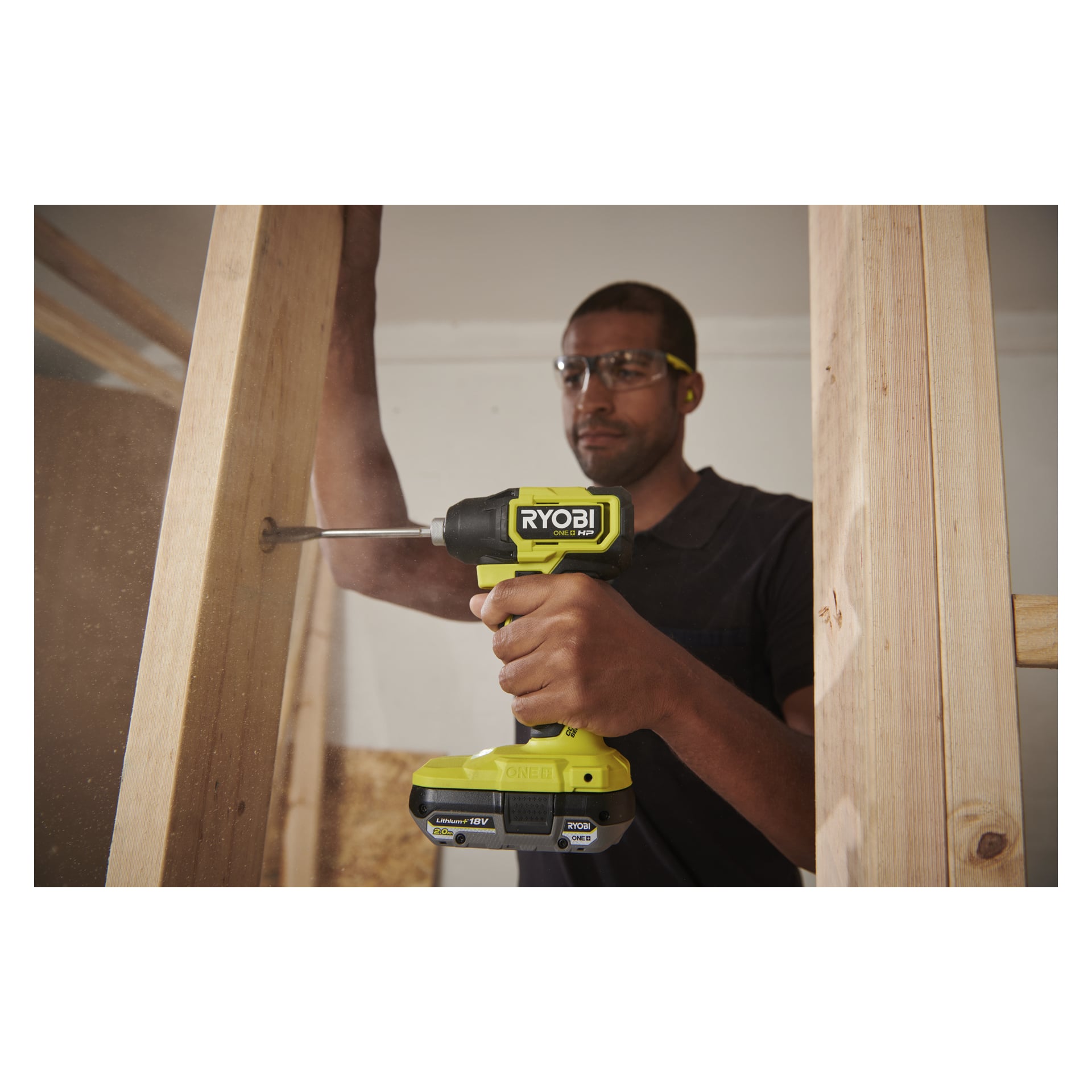 En person med skyddsglasögon slår in en skruv i en träram med hjälp av en Ryobi One+ HP Brushless slagskruvskruv, träbalkar bakom.