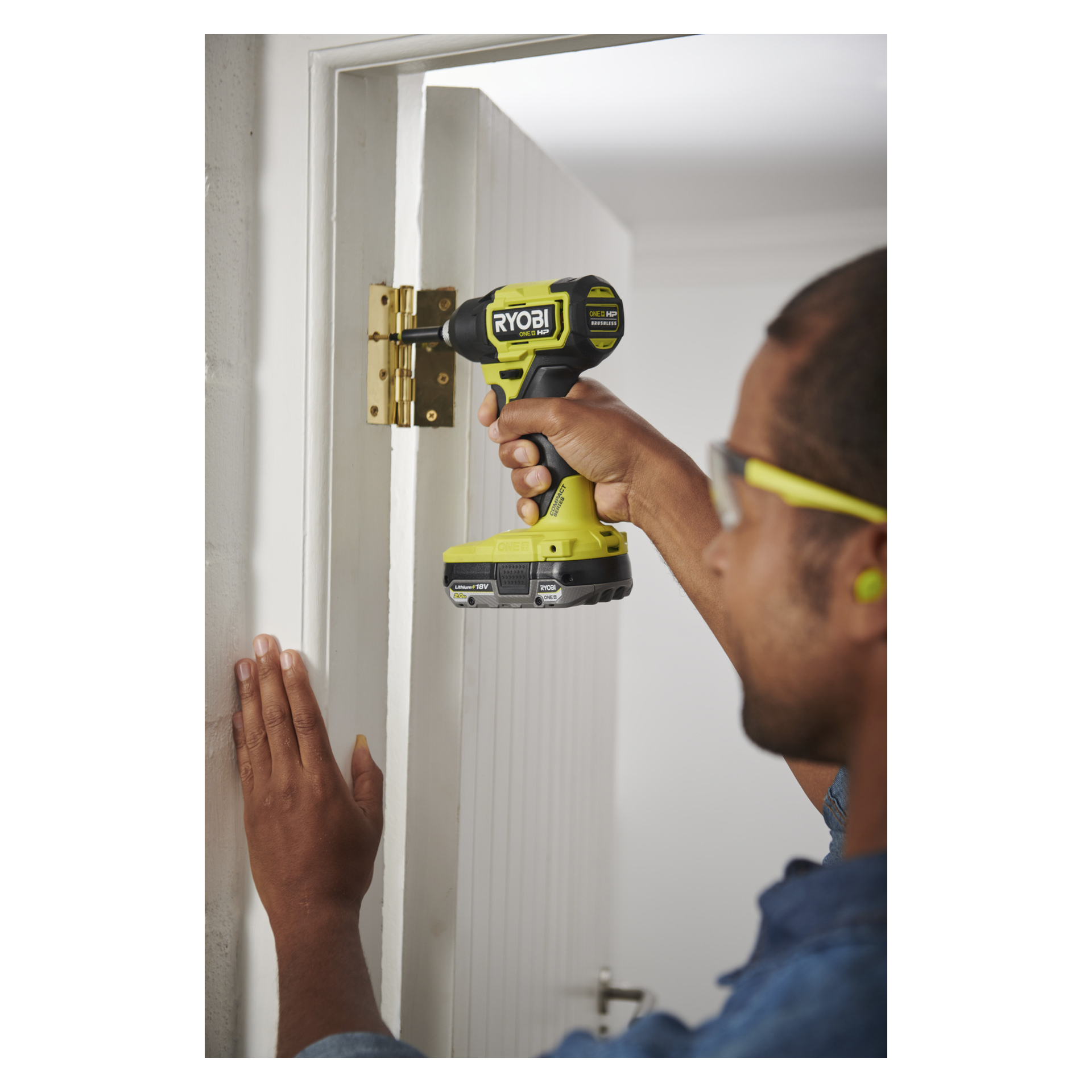 En person med skyddsglasögon och hörselskydd använder en Ryobi One+ HP slagmotor på ett gångjärn och stabiliserar en vit dörrkarm.