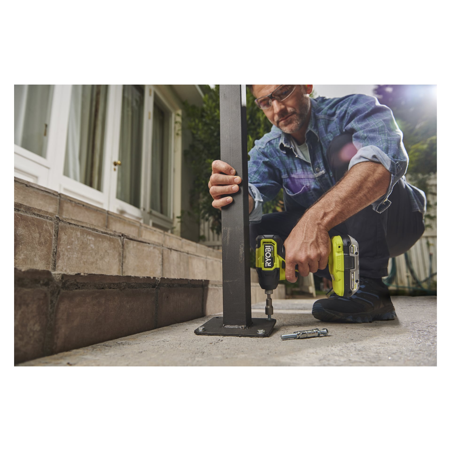 Någon använder en Ryobi One+ HP Brushless slagmotor RID18C1-0 för att fästa en metallstolpe i betong via utomhustrappor. Skyddsglasögon och skruvar finns på plats.