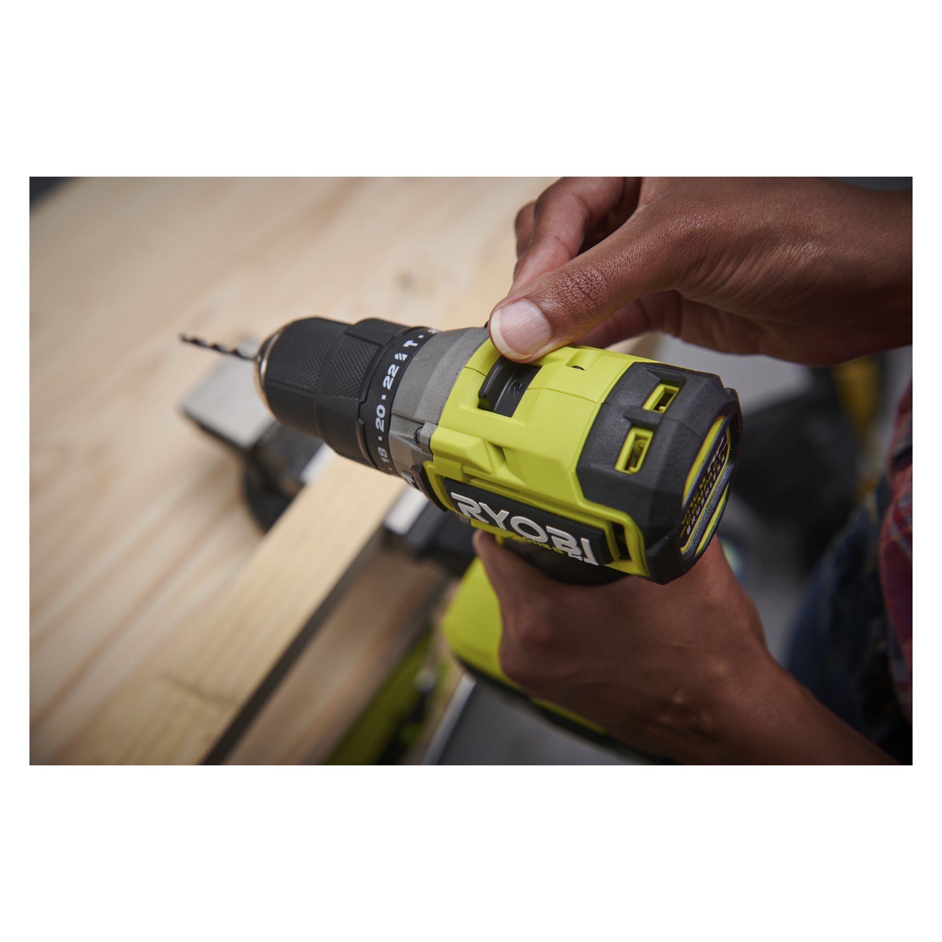 Någon justerar inställningarna på en grön och svart Ryobi One+ HP borstlös skruvmejsel och håller den över fastklämt trä på en arbetsbänk.