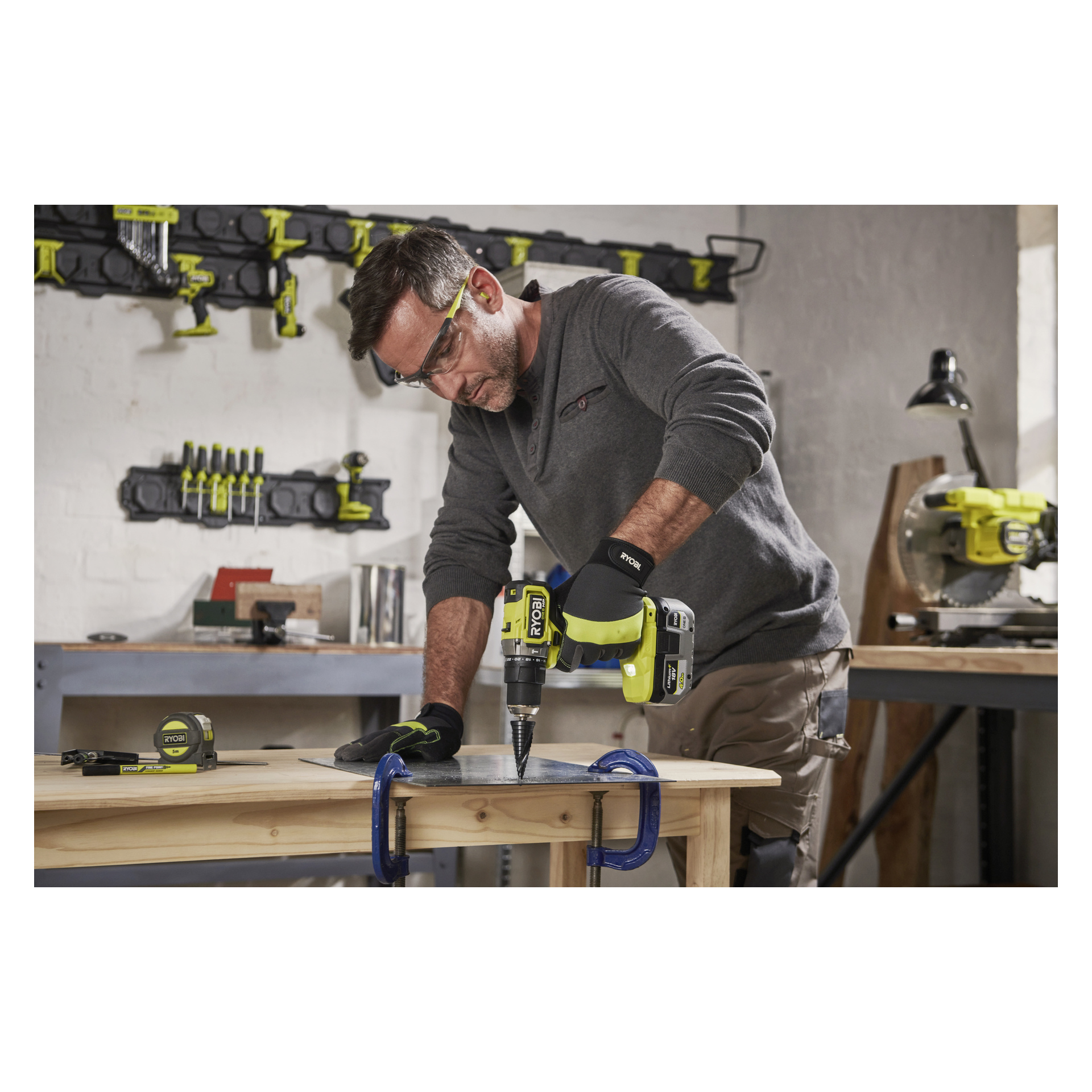 En person i skyddsutrustning borrar i en klämd metallplåt med en Ryobi One+ HP borstlös skruvmejsel i en verkstad full av verktyg.