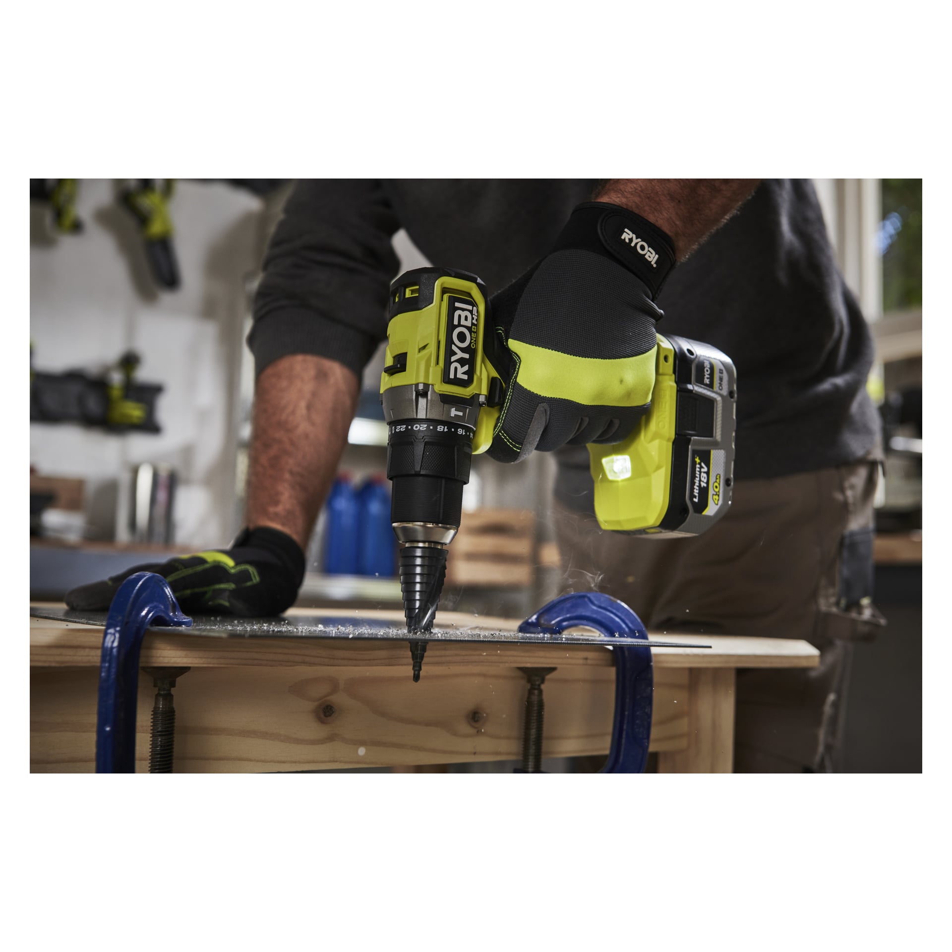 Handskbeklädda händer manövrerar en Ryobi One+ HP borstlös skruvmejsel med en stegborr, som borrar metall fastklämd på en träarbetsbänk.