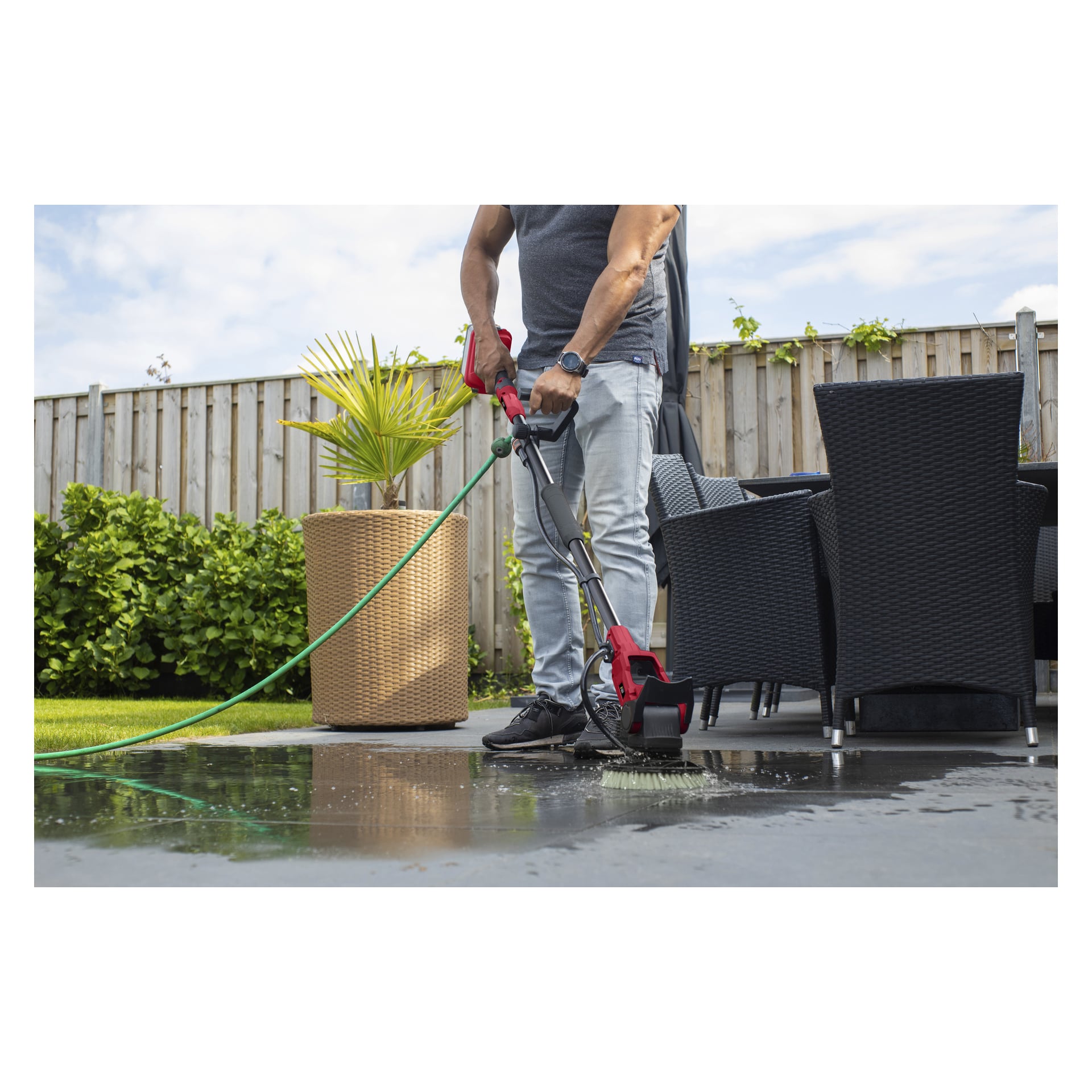 Ktoś czyści zewnętrzny taras szczotką Meec Tools PW-18V HOME 500 i zielonym wężem; krzesła, stół i roślina za nimi.