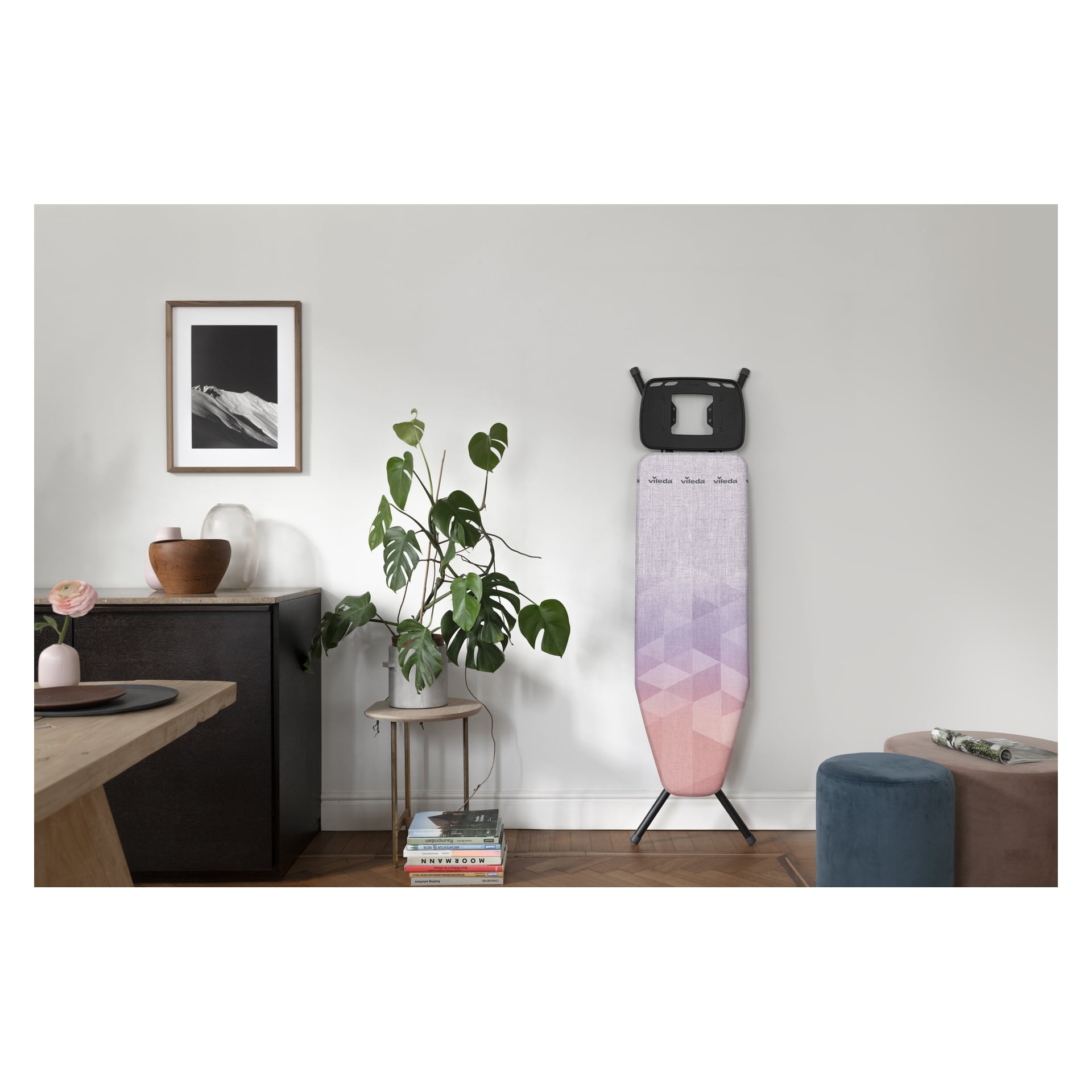 Vileda Ironing Board Diamond med rosa och lila geometriskt omslag står bredvid en grön växt, skänk, foto och böcker.