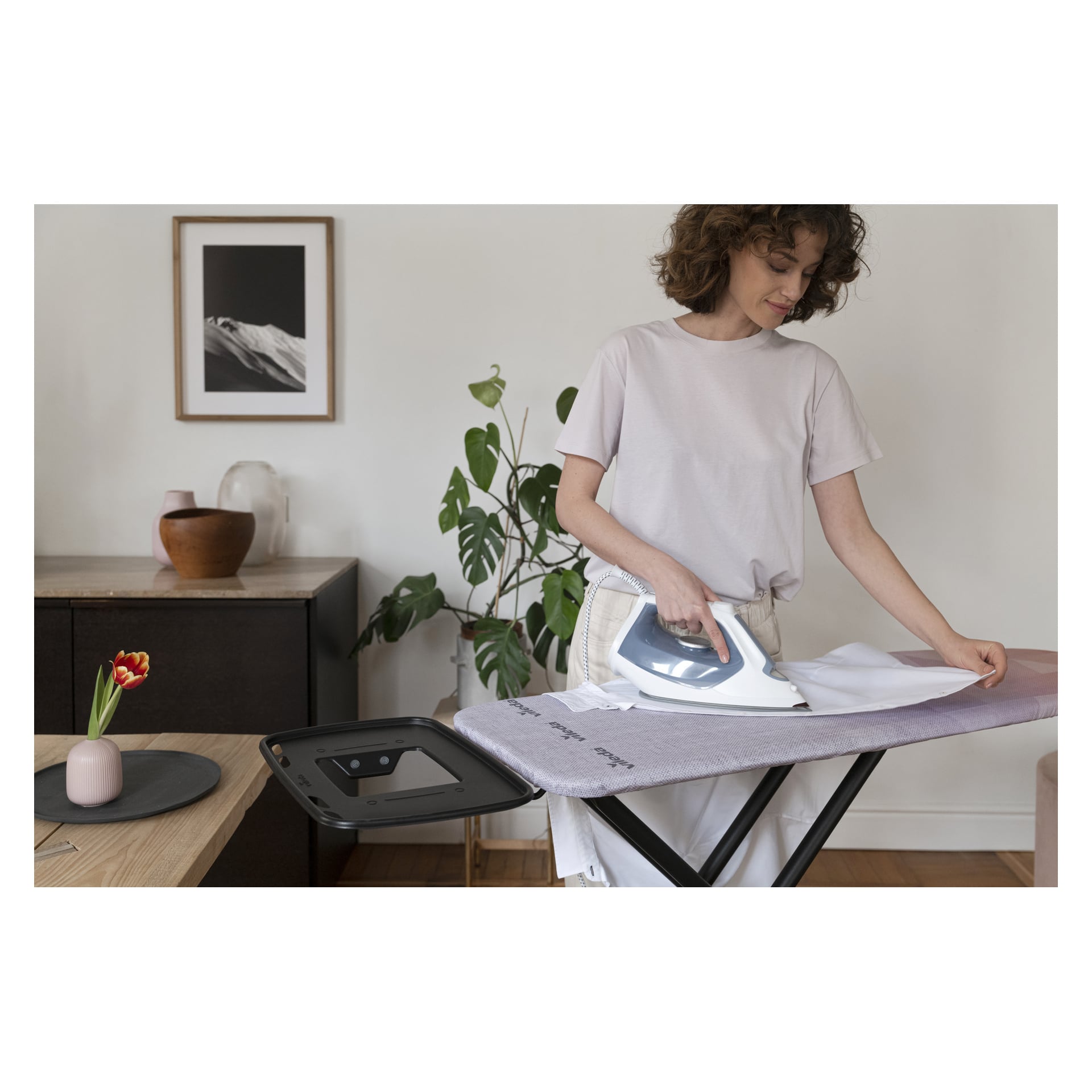 En kvinna stryker en vit skjorta på Vileda Ironing Board Diamond inomhus, med dekoration bakom och ett bord med vas och tulpan framför.