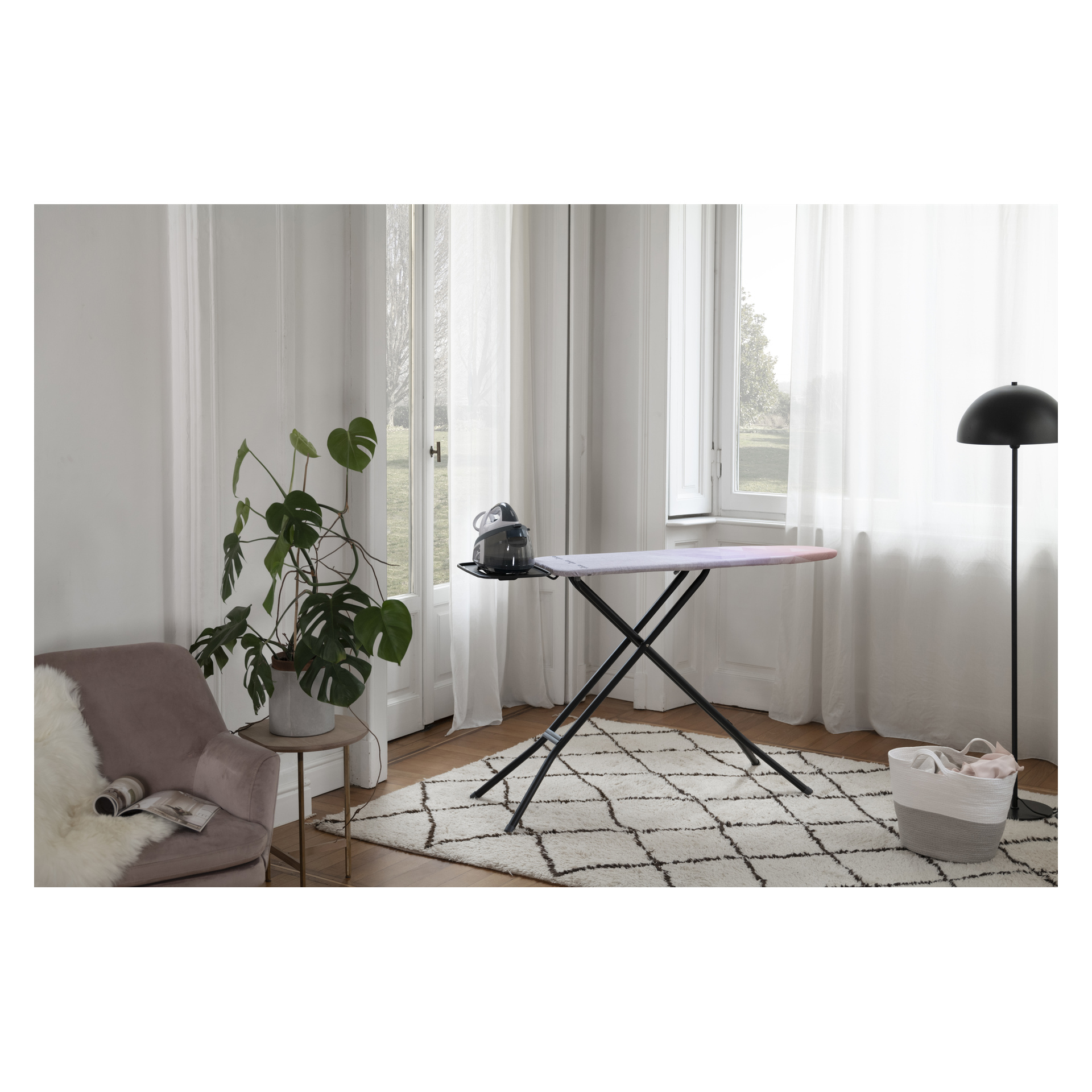 Vileda Ironing Board Diamond med strykjärn står på en matta i ett ljust rum med mysig inredning, växter och tunna gardiner.