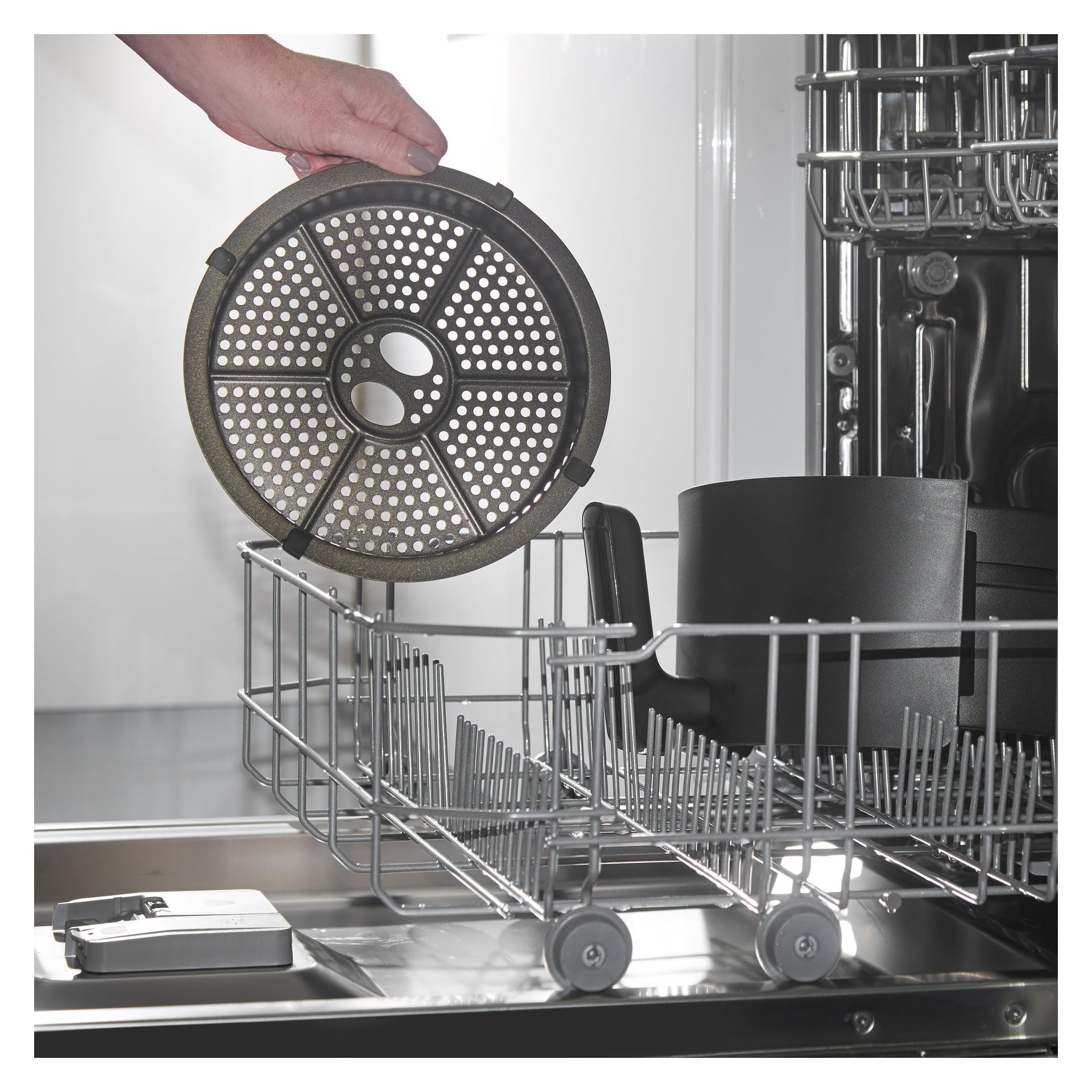 En hand laddar det runda, perforerade metallfiltret från en Russell Hobbs Satisfry Panoramic Airfryer i diskmaskinens nedre rack.