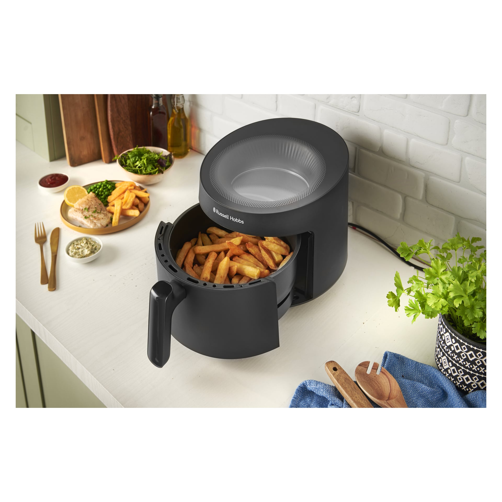 En svart Russell Hobbs Satisfry Panoramic Airfryer på köksbänken med pommes frites, omgiven av tallrikar med mat, örter och redskap.