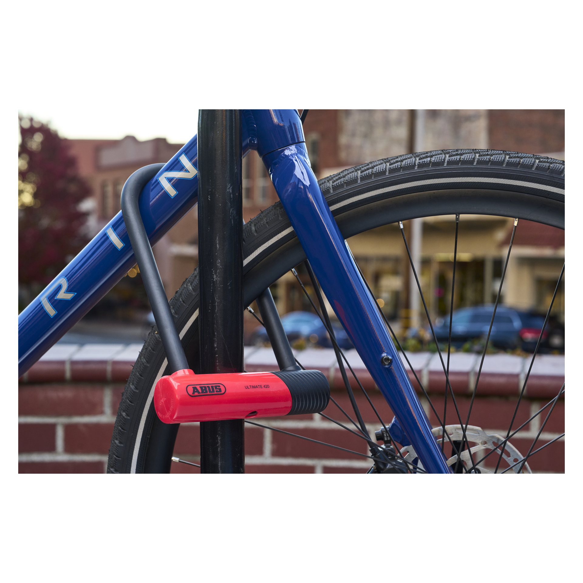 Blå cykel fastsatt i en metallställning med en röd ABUS U-lock 420K Ultimate; framhjul, ram och en suddig gata i bakgrunden.