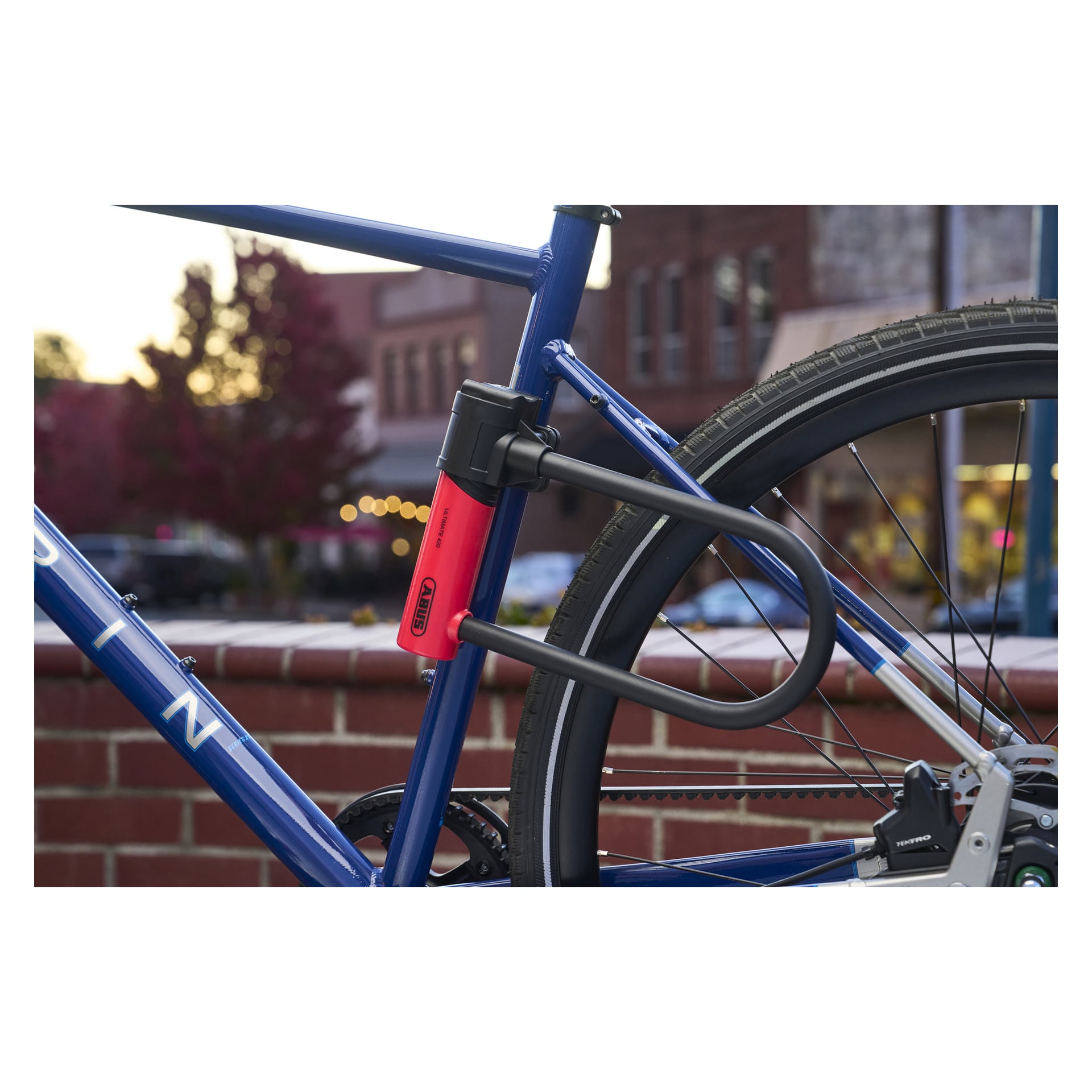 En blå cykel fastsatt med en svart och röd ABUS U-lock 420K Ultimate runt ramen och bakhjulet, lutad mot en tegelvägg utanför.