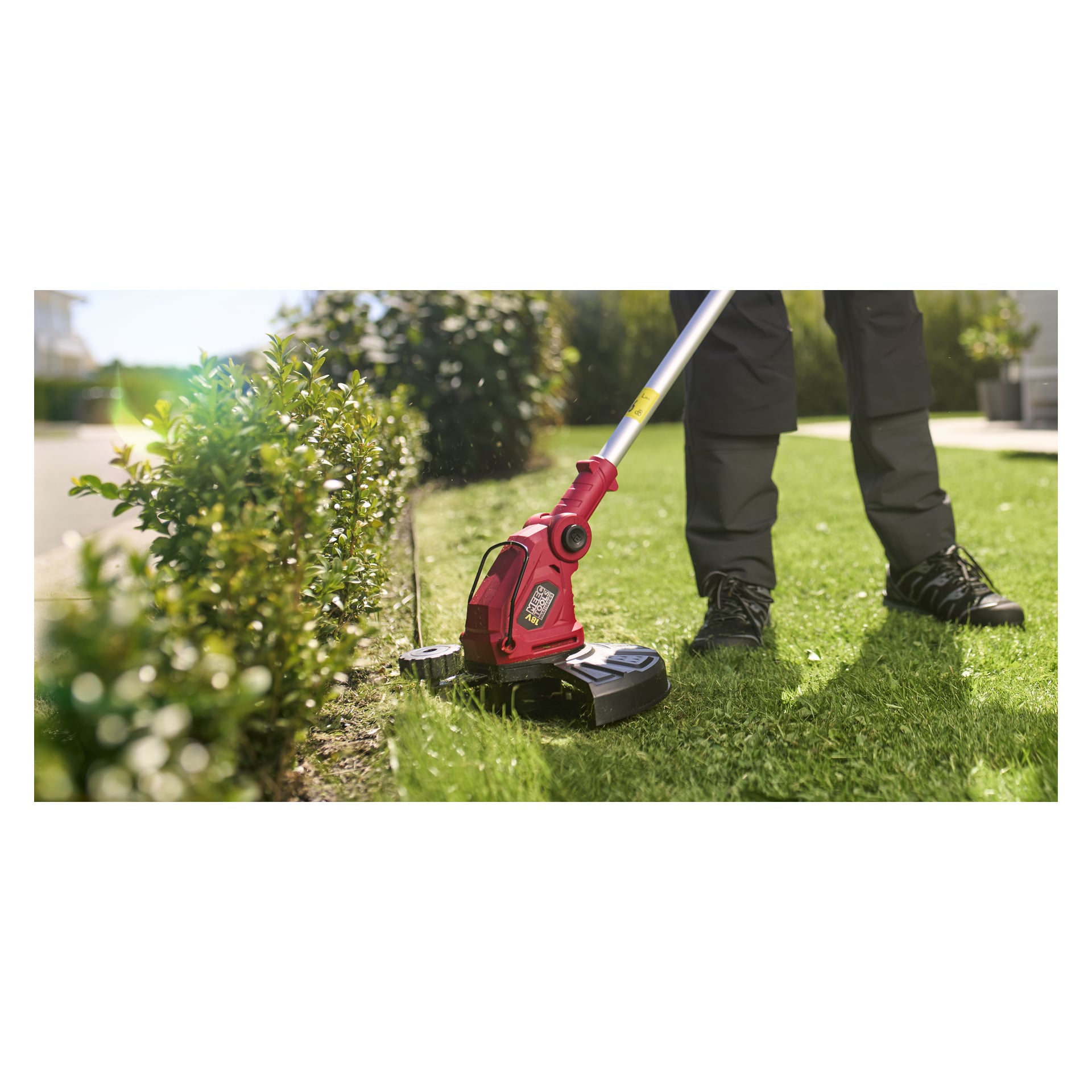 Ubrany w ciemne spodnie i buty, osoba obcinuje trawnik przy żywopłocie, używając podkaszaka Meec Tools Multiseries GT-18V HOME 500.