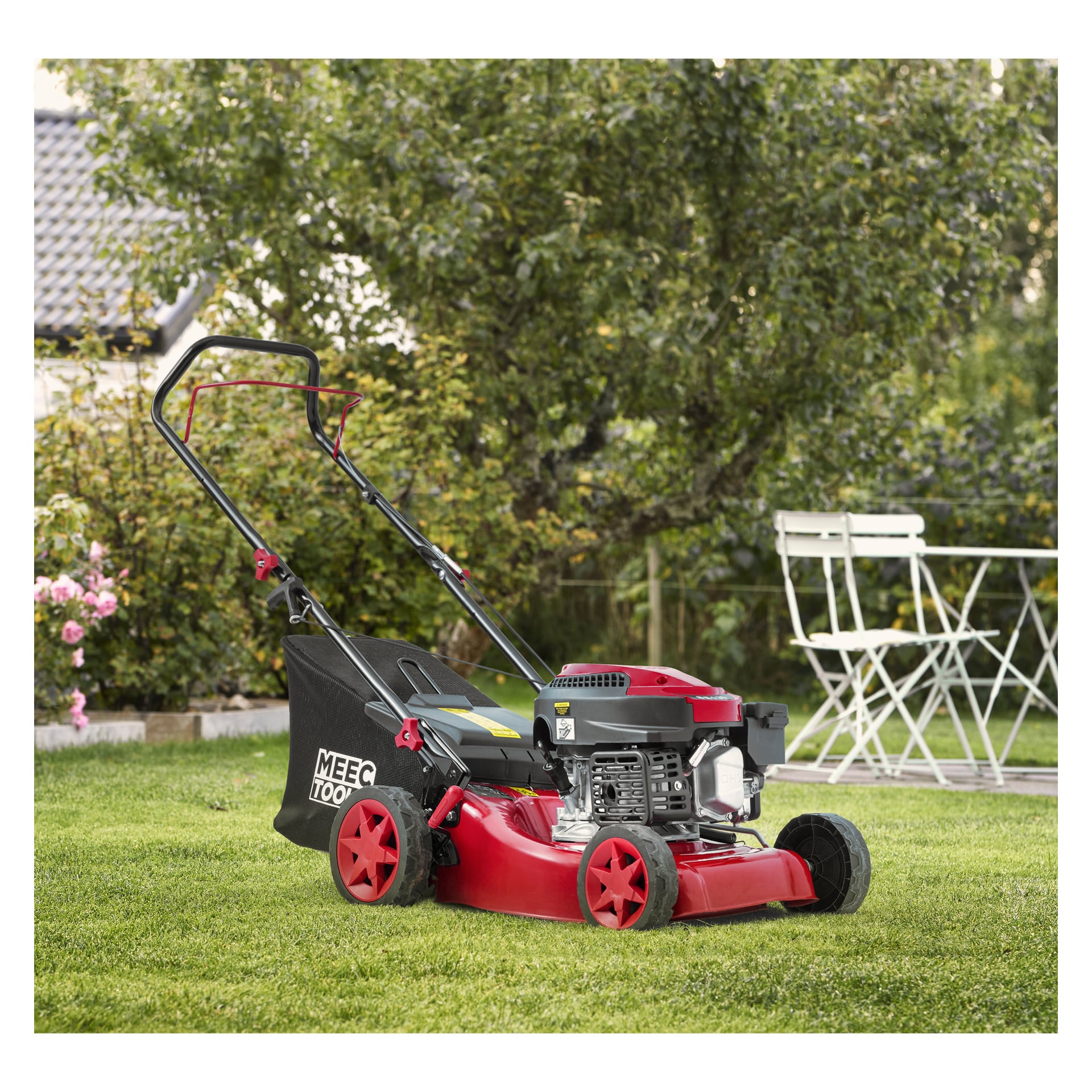En röd och svart bensingräsklippare från Meec Tools står på en gräsmatta bredvid ett vitt utebord och två stolar, omgiven av träd och blommande buskar.